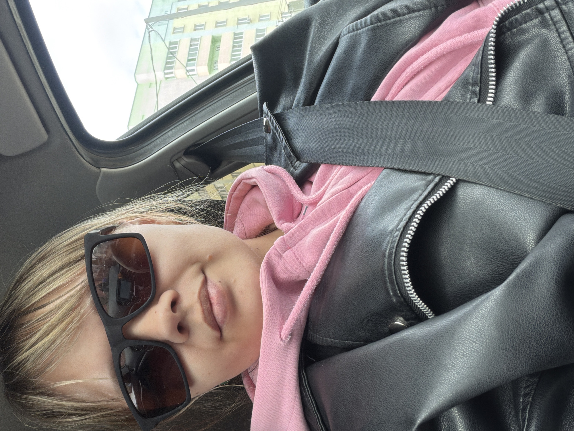 A close-up, portrait-oriented selfie shows a person with blonde hair and dark sunglasses seated inside a vehicle, likely a car. The image is rotated 90 degrees clockwise, making the person appear to be lying down with their head on the left side of the frame. The subject is wearing a pink hooded sweatshirt under a black leather jacket, with a black seatbelt fastened across their chest. Their face is partially visible, from the bridge of the nose to the chin, with lips slightly parted in a neutral or subtle expression. The sunglasses reflect some interior elements, possibly the camera used to take the photo.
The interior of the car, including part of the headliner and window frame, is visible. Through the car window and sunroof, a bright, clear sky is seen, indicating daytime. In the background, a light green and yellow building with multiple windows is visible, suggesting an urban or suburban setting. The overall mood is casual, capturing a moment during transit. There is no visible text.