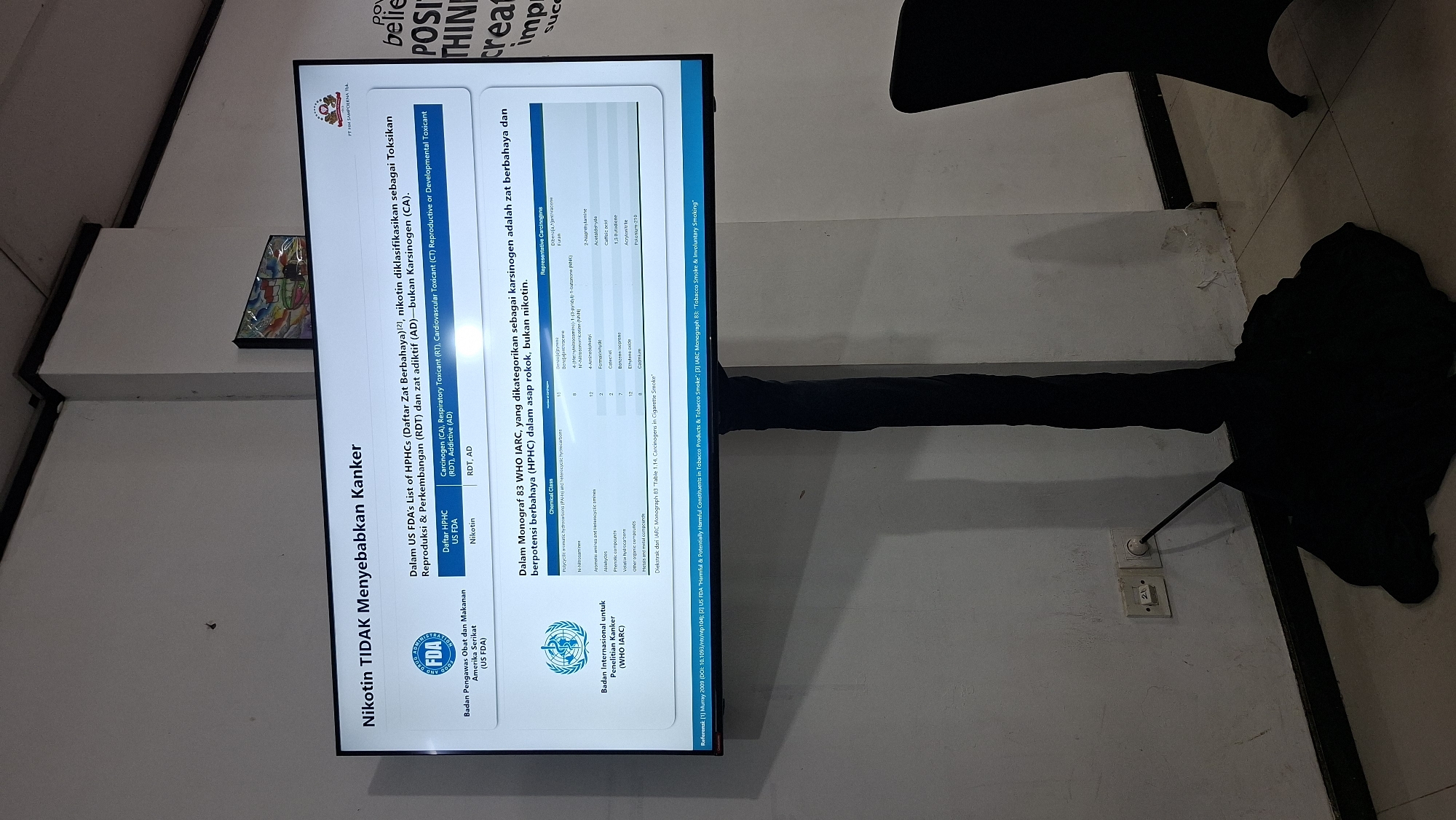 The image shows a presentation slide displayed on a large screen, likely in an indoor setting in Palembang, Indonesia. The slide's title, "Nikotin TIDAK Menyebabkan Kanker" (Nicotine Does NOT Cause Cancer), is prominently displayed. The slide contains information from the US FDA's list of HPHCs and a monograph from WHO IARC, categorizing substances as carcinogens. Text and logos from the US FDA and WHO are visible, as well as a table listing chemical classes and representative carcinogens. The screen is mounted on a black stand. In the background, a white wall with electrical outlets and a power cord is visible. The overall lighting suggests it's likely an indoor event or presentation.