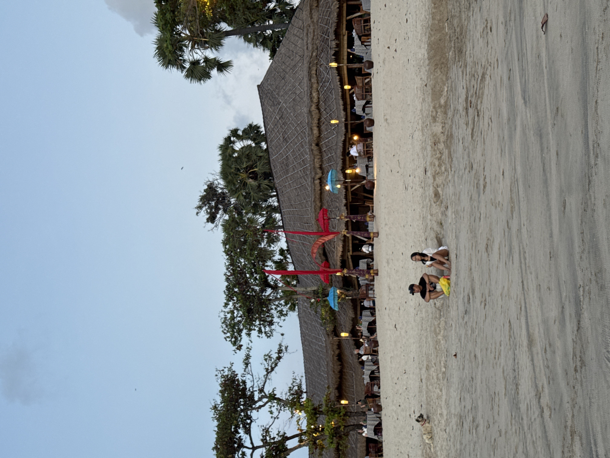 The image shows a beach scene in Kuta Selatan, Indonesia. Along the edge of the beach is a thatched-roof structure, possibly a restaurant or bar, with numerous people seated at tables inside. The beach itself appears sandy, with two people crouching or sitting on the sand. One person is wearing a white shirt and the other a black shirt. A dog is visible a short distance away. The sky is overcast. Trees are interspersed along the edge of the beach near the structure.