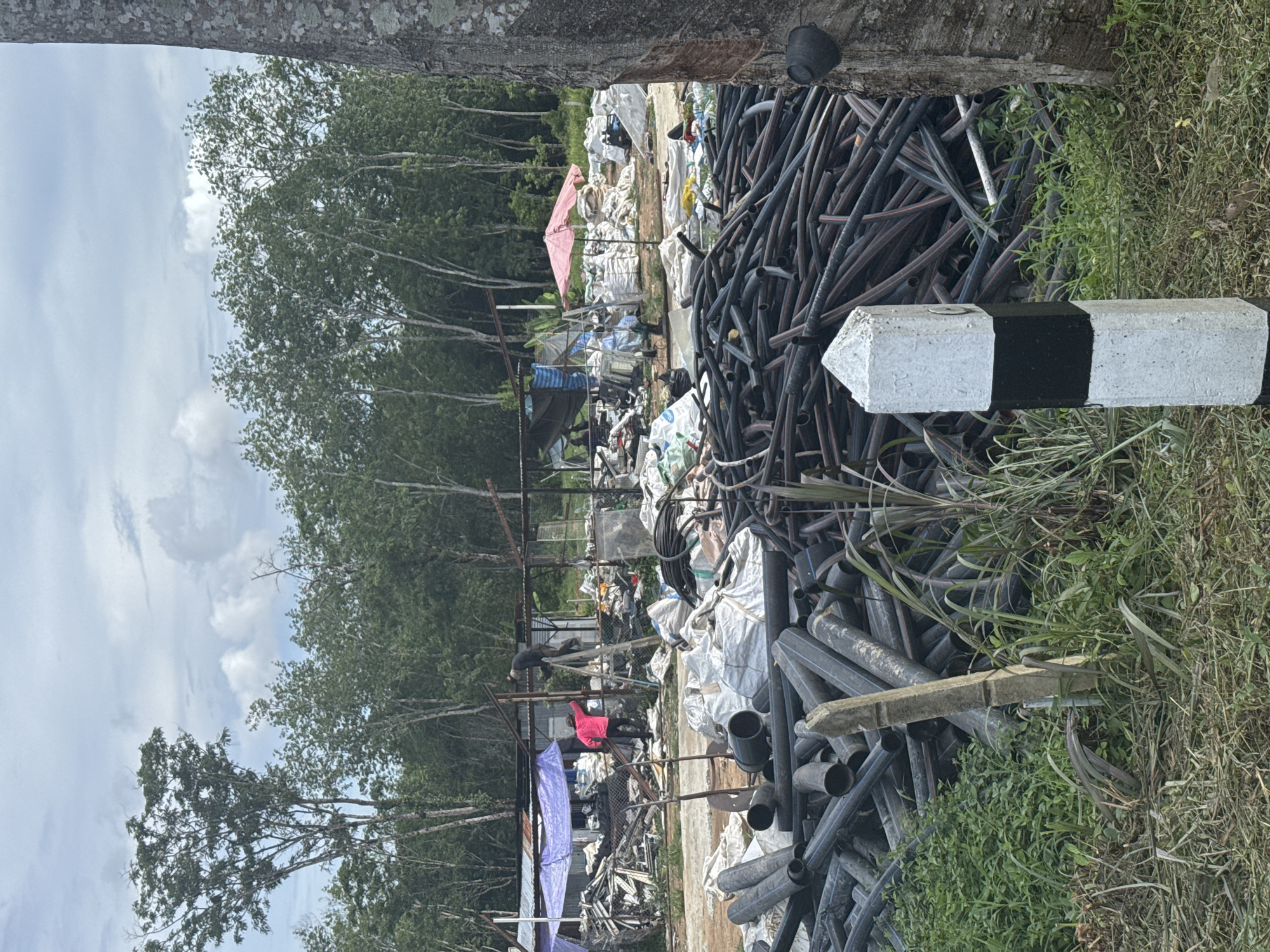 The image depicts a scene in Pa Khlok, Thailand, characterized by environmental pollution and waste accumulation. A large pile of black plastic pipes dominates the foreground, partially obscuring a roadside marker painted with alternating white and black sections. Behind the pipes, a cluttered area with makeshift structures and piles of discarded materials is visible.
Two people are present in the scene, one wearing a red shirt and the other in black, both appearing to be working amidst the debris. The background features dense vegetation, including tall trees with green foliage, indicative of a tropical environment. The sky is overcast, with clouds covering most of the visible area. Overall, the image conveys a sense of disorder, environmental degradation, and human activity focused on waste management or collection in an informal setting.