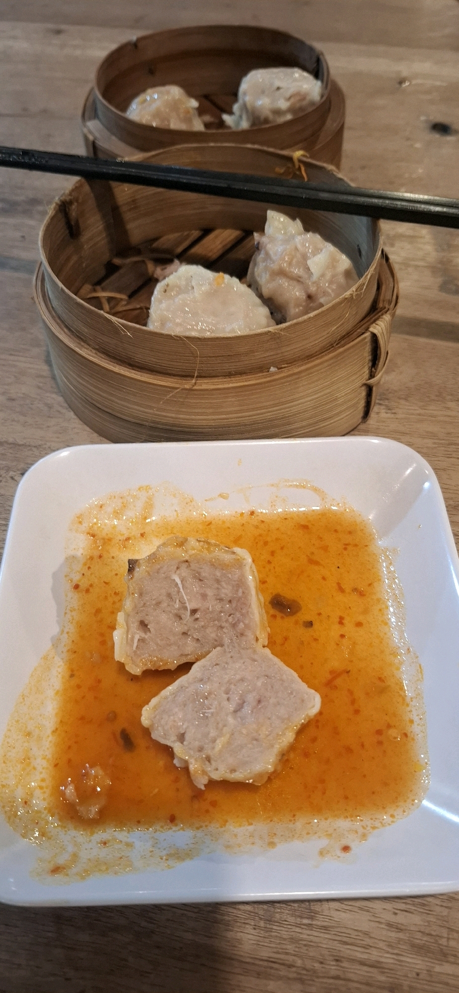 Two bamboo steamers filled with dim sum sit on a wooden table. The steamers are stacked one above the other, with chopsticks resting across the top steamer. In the foreground, a white plate holds two pieces of dim sum that appear to have been cut and are partially submerged in an orange sauce. The table surface is made of light-colored wood planks. The scene is lit by overhead lighting, casting soft shadows. The overall impression is one of a meal being served or enjoyed.
