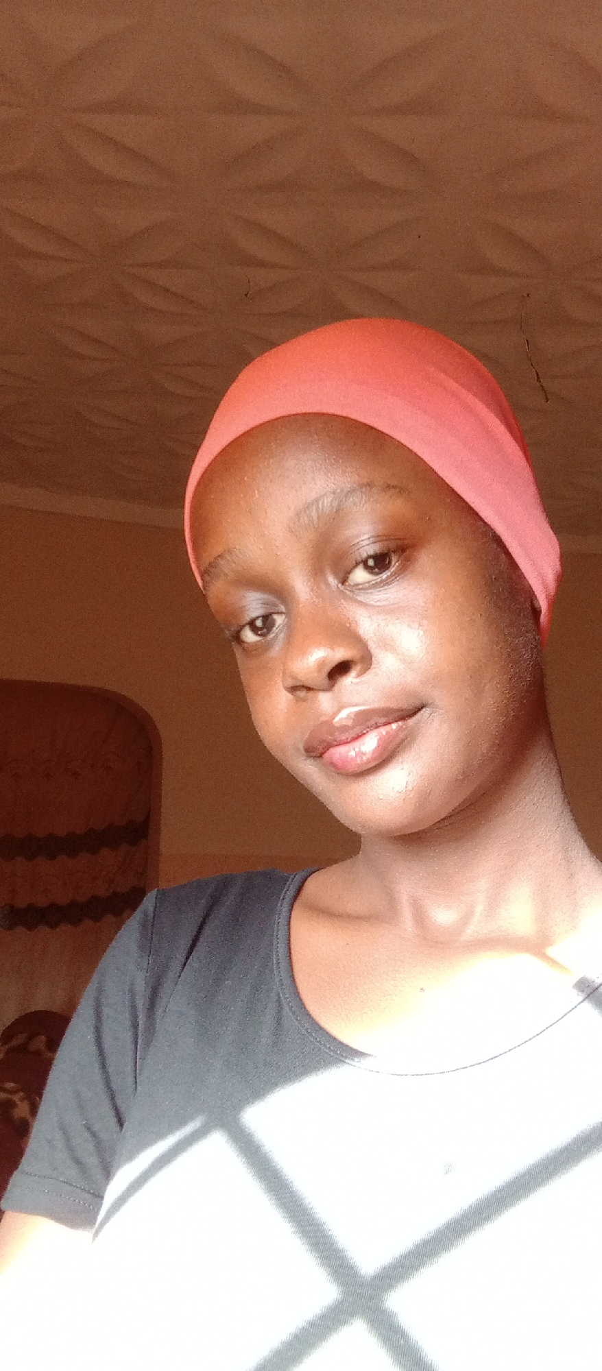 A close-up selfie of a young woman with dark skin, wearing a coral-colored headscarf and a black t-shirt. Her lips are glossy, and she is looking slightly upwards towards the camera. The lighting suggests an indoor setting. The background includes a textured wall and a chair. The specific location, Akwanga, Nigeria, is not ascertainable from the visual data.