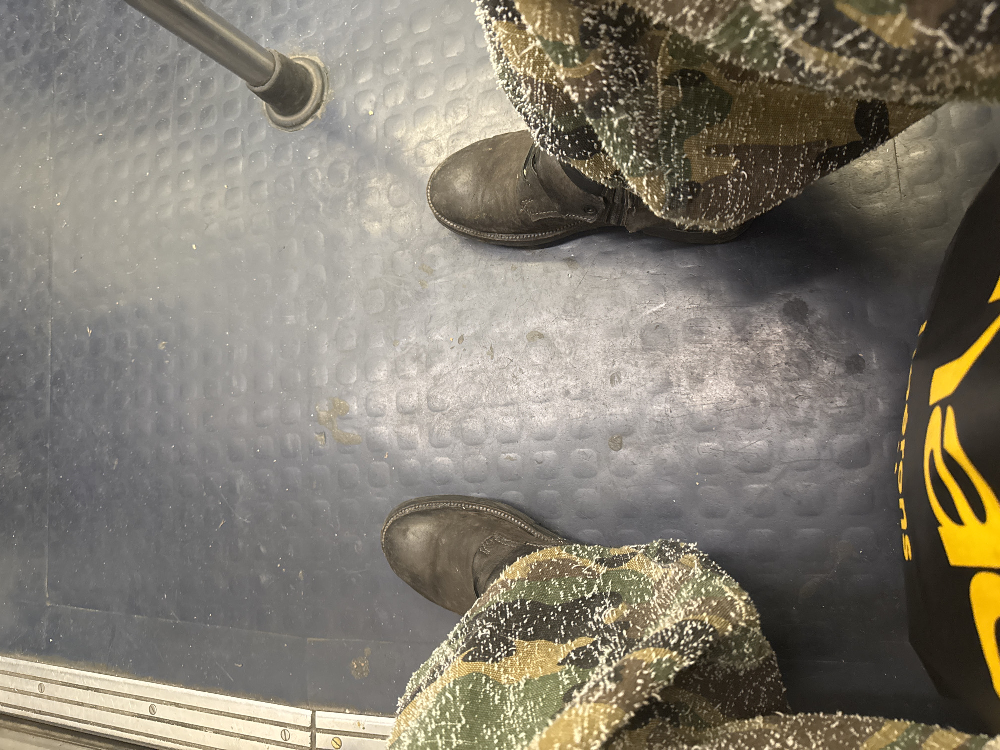 The image shows a view downward onto the floor of a public transportation vehicle, potentially a train or bus, identifiable by the textured dark gray floor and metallic railing running along the bottom edge of the frame. A vertical metal pole with a rubber base is visible in the upper left quadrant. Two boots are present, both appearing to be dark-colored leather work boots. The boots are worn with camouflage pants that have a speckled, white appearance, suggesting possible contact with snow or a similar substance. The right side of the image includes a partial view of a black bag with large yellow graphics. The location is unknown.