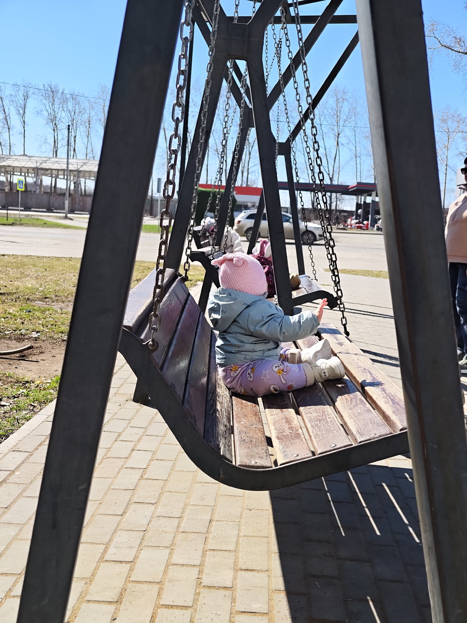 A young child sits on a wooden bench swing in Chaikovsky, Russia. The child, wearing a pink hat, light blue jacket, and patterned pants, is facing away from the viewer with their right arm extended. Other people, including another child, are partially visible on the swing and nearby. The swing is suspended by metal chains from a dark metal frame. The ground is paved with rectangular blocks. In the background, there is a building structure, bare trees, and a gas station. The sky is blue and clear.
