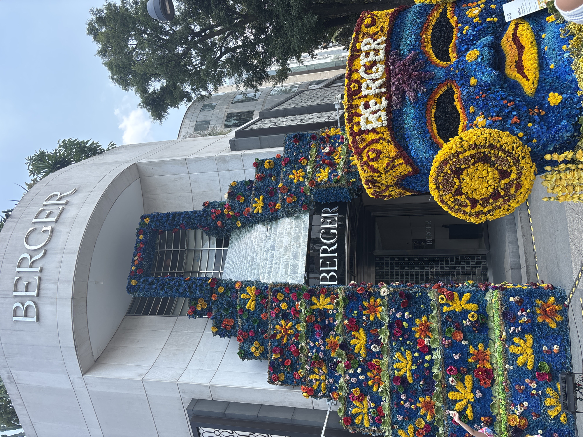 The image captures the façade of a "BERGER" store in Mexico City, Mexico, adorned with an elaborate floral display. The store itself is a modern building with a curved corner and a prominent "BERGER" sign above the entrance. The floral arrangement, executed predominantly in shades of blue, yellow, and red, covers the side of the building in a stepped pattern. A large floral mask with the word "BERGER" above it is positioned to the right of the store's entrance. The sky is partially visible in the upper left of the frame, along with parts of a tree and buildings in the background. There is a glimpse of a person's hand in the bottom right, suggesting the presence of onlookers. The image conveys a sense of a grand, temporary floral installation at a retail location.