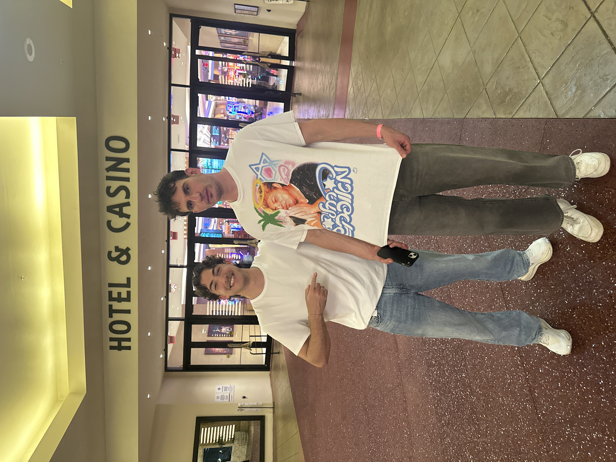 The image shows two males standing in front of the entrance to the Hollywood Seminole Hotel & Casino. The male in the foreground is wearing a white t-shirt and blue jeans, and is smiling. The male behind him is wearing a white t-shirt with a graphic design on the front and grey pants. The location is the Hollywood Seminole Reservation, United States of America.