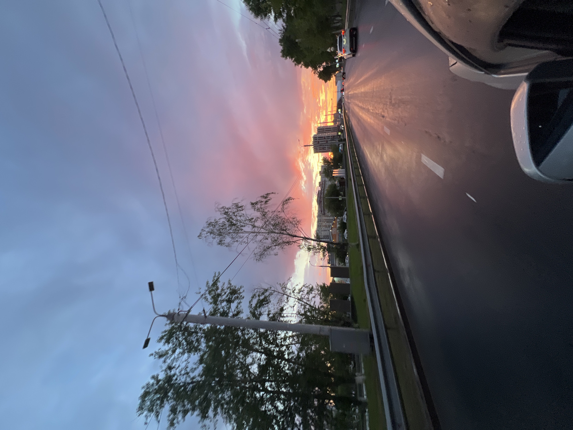 The image depicts a sunset scene on a city street in Irkutsk, Russia. The sky is a dramatic display of pink, orange, and purple hues, with clouds reflecting the vibrant colors. A wet asphalt road stretches forward, with lane markings visible. The wet surface of the road reflects the colorful sky, creating a shimmering effect. Several cars are visible on the road, suggesting ongoing traffic. Trees line the left side of the road, with a streetlamp and a utility box next to them. Buildings, including what appear to be apartment complexes, are visible in the distance on the right side of the road, indicating an urban setting. The perspective suggests the photo was taken from inside a vehicle, with the side mirror and part of the car visible in the foreground.