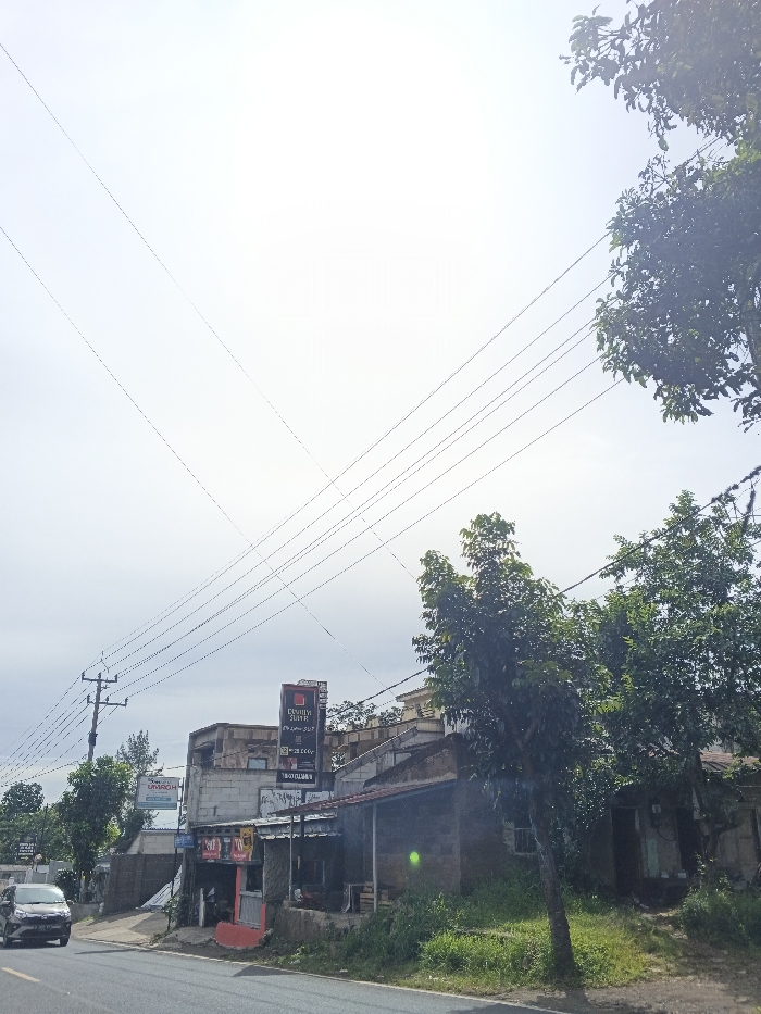 A street scene features a small, weathered building with a red sign and a larger sign above it. Power lines crisscross overhead against a bright sky. Green trees frame the right side of the image. A car drives along the asphalt road in the lower-left corner. The scene conveys a sense of rural or suburban simplicity.
