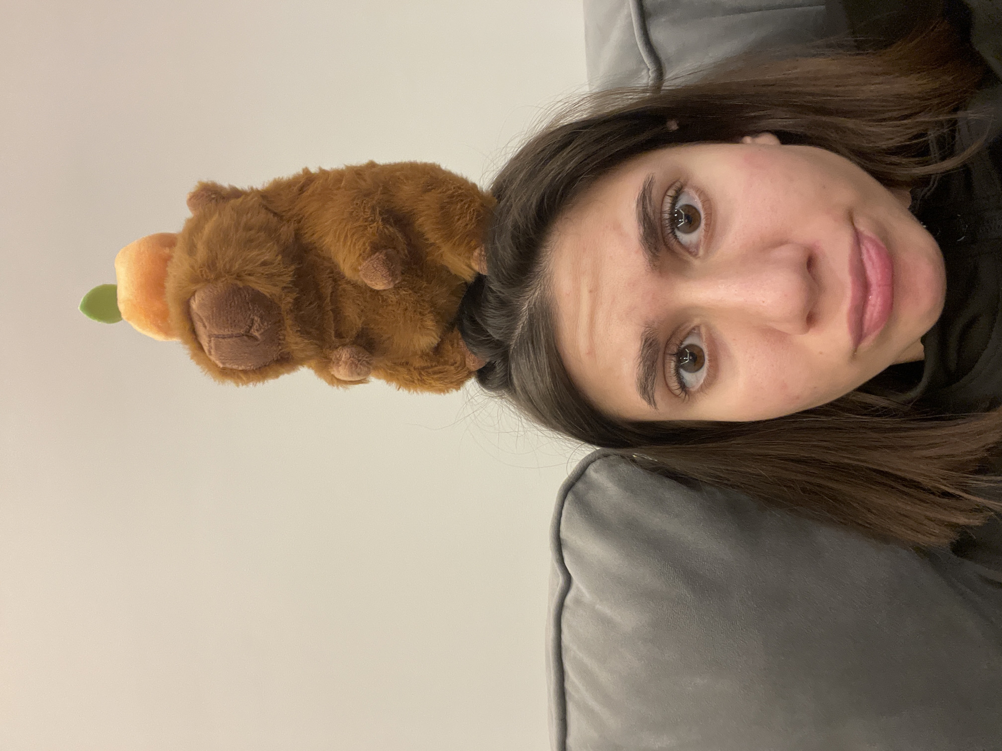 A person is laying on a gray couch with their head near the top right corner of the image. There is a brown stuffed animal of what looks like a platypus sitting on their head. The platypus has an orange and green object near its face. The background is a plain white wall. The image does not contain any information that indicates the location in the city of Sofia, Bulgaria.