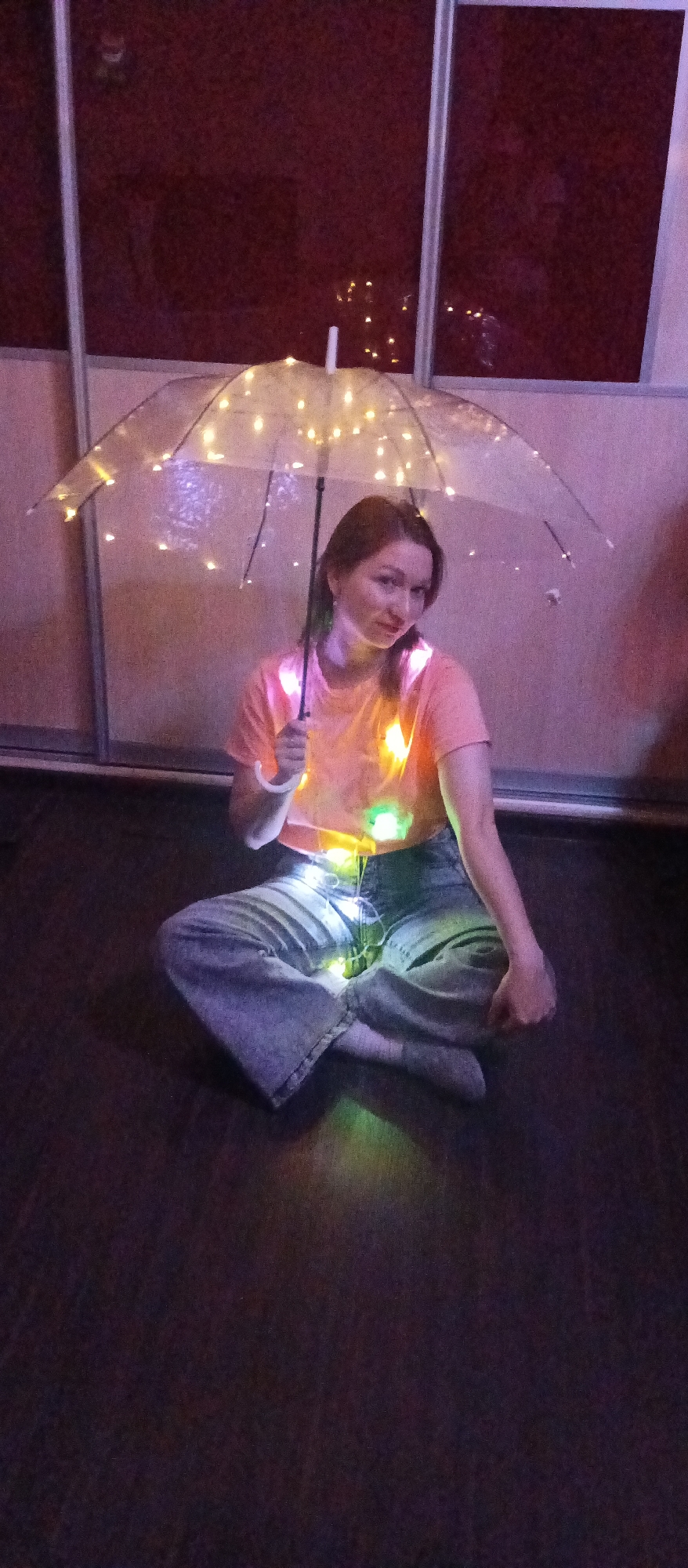 A young woman is seated on a dark floor, cross-legged, holding a clear umbrella adorned with warm white fairy lights. She wears a short-sleeved, light orange or peach t-shirt and light blue, loose-fitting jeans, with light-colored socks or barefoot. Additional fairy lights, including some colorful ones (green, yellow, pink), are draped around her chest and lap, contributing to the glowing effect.
The woman looks directly at the camera with a gentle expression and a slight smile. Her hair is dark brown and appears to be pulled back loosely. The background consists of dark panels, possibly a closet door or wall, framed by light-colored vertical and horizontal strips. Reflections of the fairy lights and faint outlines of the woman's figure are visible on these dark panels.
The overall scene is dimly lit, suggesting an indoor setting in the evening or night, with the primary illumination coming from the numerous fairy lights on the umbrella and around her body. The image has a warm, ethereal glow created by the lights, contrasting with the dark floor and background. The image exhibits some graininess. No visible text is present.