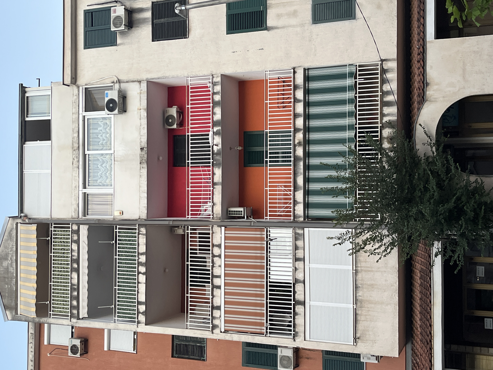 The image shows a close-up, eye-level view of a multi-story apartment building in Budva, Montenegro, under a clear sky. The building has a textured, off-white facade and features multiple balconies with white railings. The balconies are equipped with various amenities, including retractable awnings in striped patterns of yellow, grey, and orange, as well as green and white stripes. Several windows are visible, some with closed green shutters, and others with white blinds. Air conditioning units are mounted on the exterior walls of several apartments. The building's lower section features a tiled roof and an arched entryway, suggesting it may house shops or businesses on the ground floor. A small, leafy green tree is positioned in the foreground, partially obscuring the lower part of the building. The overall scene is typical of a residential and commercial area in a European coastal city.