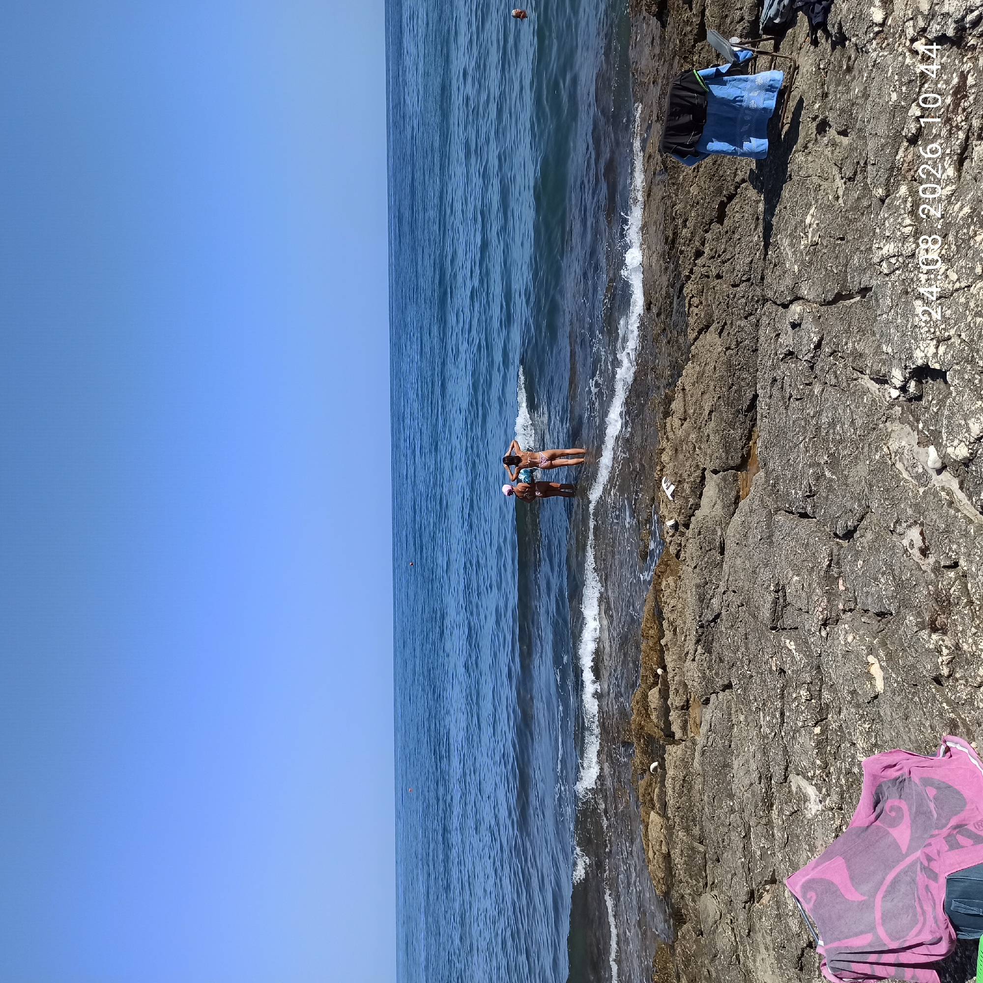 The image depicts a rocky shoreline with the Mediterranean Sea stretching to the horizon under a clear blue sky. In the foreground, a pink towel is draped over a dark bag on a rough, gray rock surface. To the right, a blue folding chair holds a blue and black garment. Further along the shore, two individuals are partially submerged in the shallow water, facing away from the camera. The sea displays gentle waves breaking on the rocks. In the distance, a single orange marker floats on the water. The scene is dated 24/08/2026 at 10:44. This location is consistent with the coastal areas of Molfetta, Italy, known for its rocky shores and proximity to the Adriatic Sea.