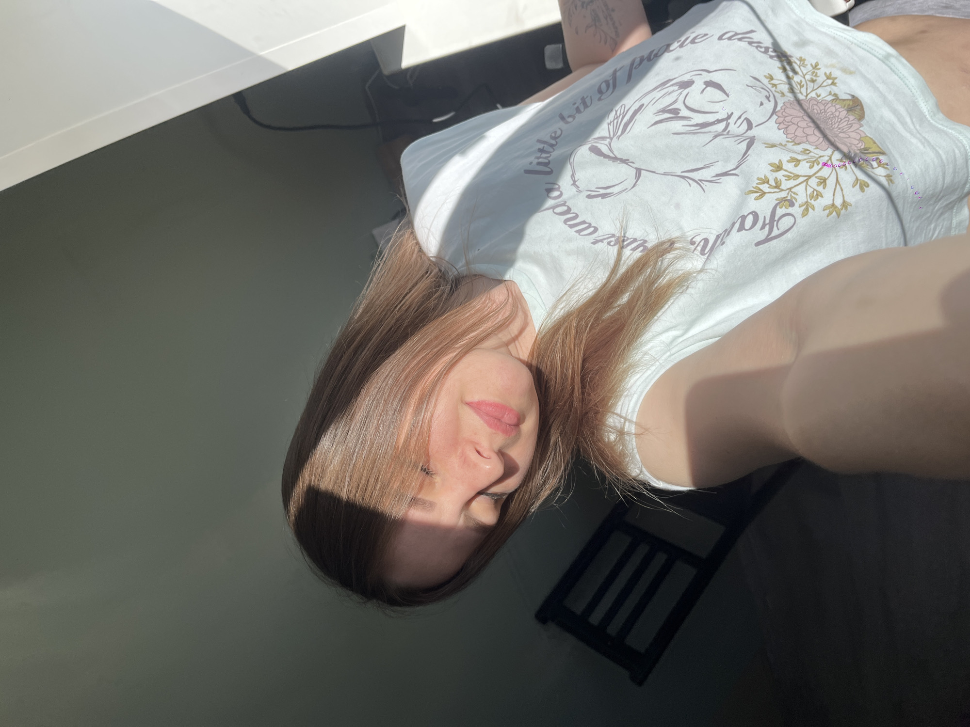 A woman is sitting in a room with sunlight, wearing a t-shirt with an image of a flower and words on it. There is a chair behind her, and a desk on the left. The location cannot be determined from the image.