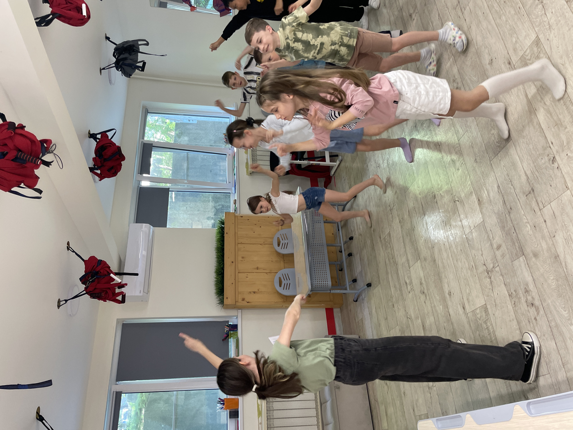 This is an indoor photograph capturing a group of children and an adult engaged in a coordinated physical activity or game. The image is oriented vertically, suggesting a rotation from its original landscape perspective.
The scene takes place in a brightly lit room with light-colored wooden or laminate flooring and white walls. Large windows allow natural light to stream in, revealing greenery and other buildings outside, indicating it is daytime. An air conditioning unit is mounted high on one wall. Several red and one grey backpack hang on hooks on the walls. In the middle ground, a metal table with two grey chairs is visible, placed beside a wooden planter box filled with artificial grass.
Approximately eight to nine individuals are visible, mostly children appearing to be between 6 and 12 years old, along with one adult. The adult, a person with long brown hair tied in a ponytail, wearing a green t-shirt and dark pants, stands towards the foreground (bottom left of the rotated image) with arms outstretched, seemingly leading the activity. The children are positioned behind the adult, mirroring the pose with their arms extended sideways, as if balancing or pretending to fly. Their expressions appear engaged and focused. Some children are barefoot, while others wear socks or shoes. The activity suggests an exercise class, a game, or a creative movement session.
No specific weather conditions are discernible, other than the general daylight. The location context provided is Sochi, Russia. There is no clearly legible text visible within the image.