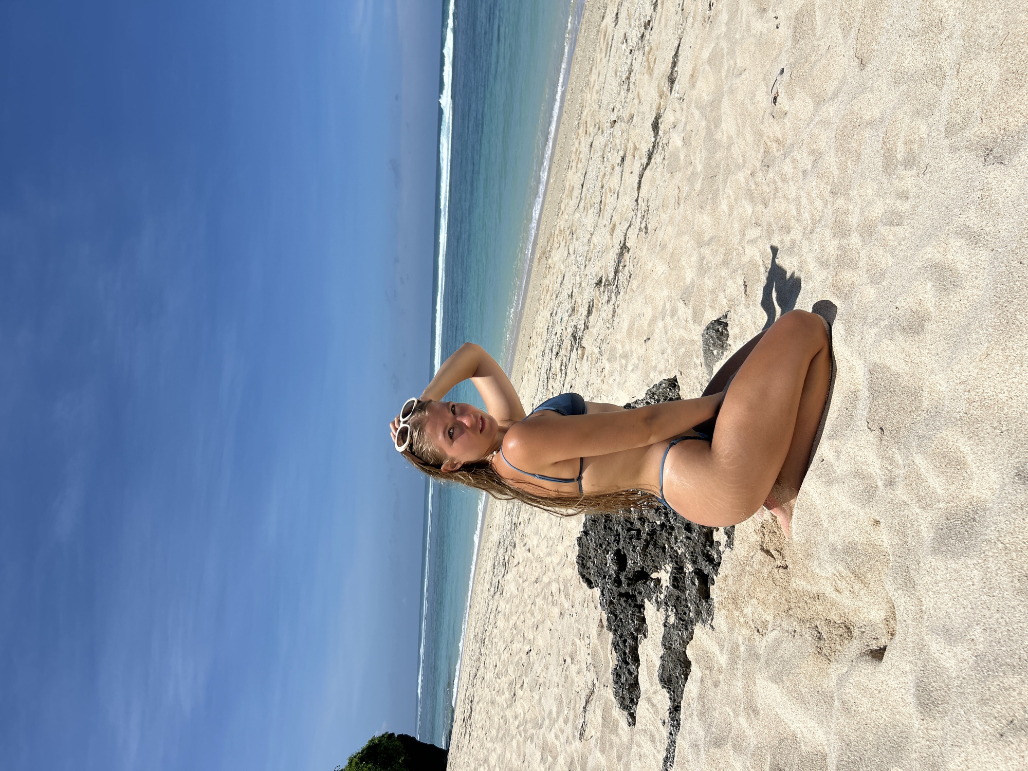 The image captures a woman in a blue bikini and sunglasses, seated on a sandy beach adjacent to a dark, rocky formation. She is holding her hair with one hand, posing for the camera. The sky is clear and blue, and the ocean with a visible wave break is in the background. The scene is located on a beach within the city of Kuta Selatan, Indonesia.