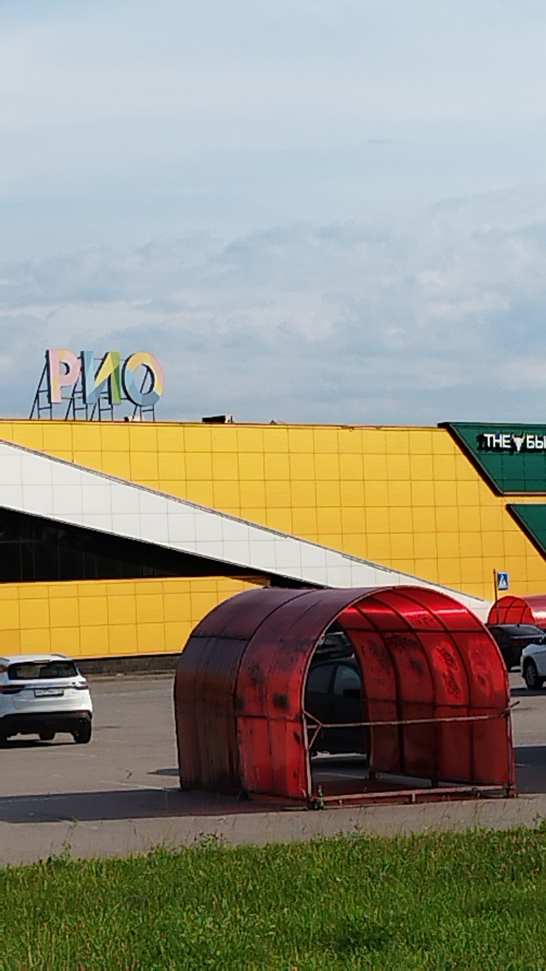 The image shows the exterior of a large commercial building, likely a shopping mall, with a yellow facade. On the roof, a colorful sign reads "РИО" in large, stylized letters. To the right, a green section of the building has a sign with white text that appears to read "THE Бык". In the foreground, there is a red, translucent tunnel-like structure, possibly a covered walkway or a car wash bay. A white SUV is parked on the asphalt in front of the red structure, and another car is visible further to the right. The ground is paved, with a strip of green grass at the bottom of the frame. The sky is partly cloudy, suggesting it is daytime. The scene appears to be a typical day at a commercial establishment. The overall impression is one of a functional, modern retail environment.