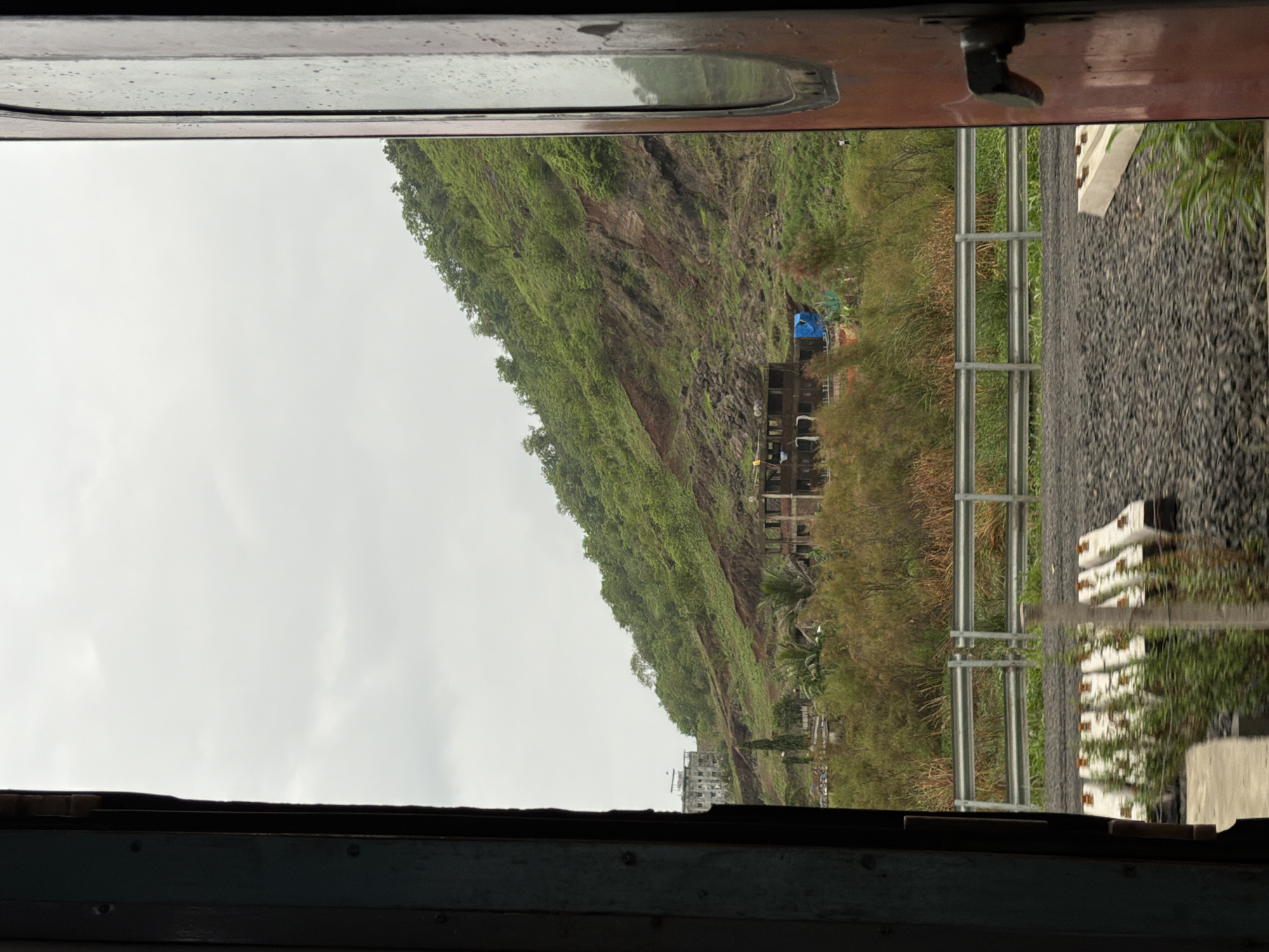 The image, taken from inside a train carriage looking out through an open doorway, displays a cloudy sky over a lush green, heavily vegetated hillside. The hillside shows signs of a landslide with exposed reddish-brown earth and rocks. At the base of the hill, partially obscured by trees and vegetation, are several unfinished or abandoned construction sites with exposed concrete structures. In the mid-ground, a metal guardrail runs parallel to a railway track, which is laid on a bed of dark gravel. In the foreground, several concrete railway sleepers are visible on the gravel. The doorway frames the view on both the left and right sides, with the left side showing a dark, weathered wooden frame and the right side a reddish-brown door with a handle. The overall scene suggests a rural or semi-rural setting near the outskirts of Vasai-Virar, India, possibly during or after monsoon season, given the greenery and the landslide.