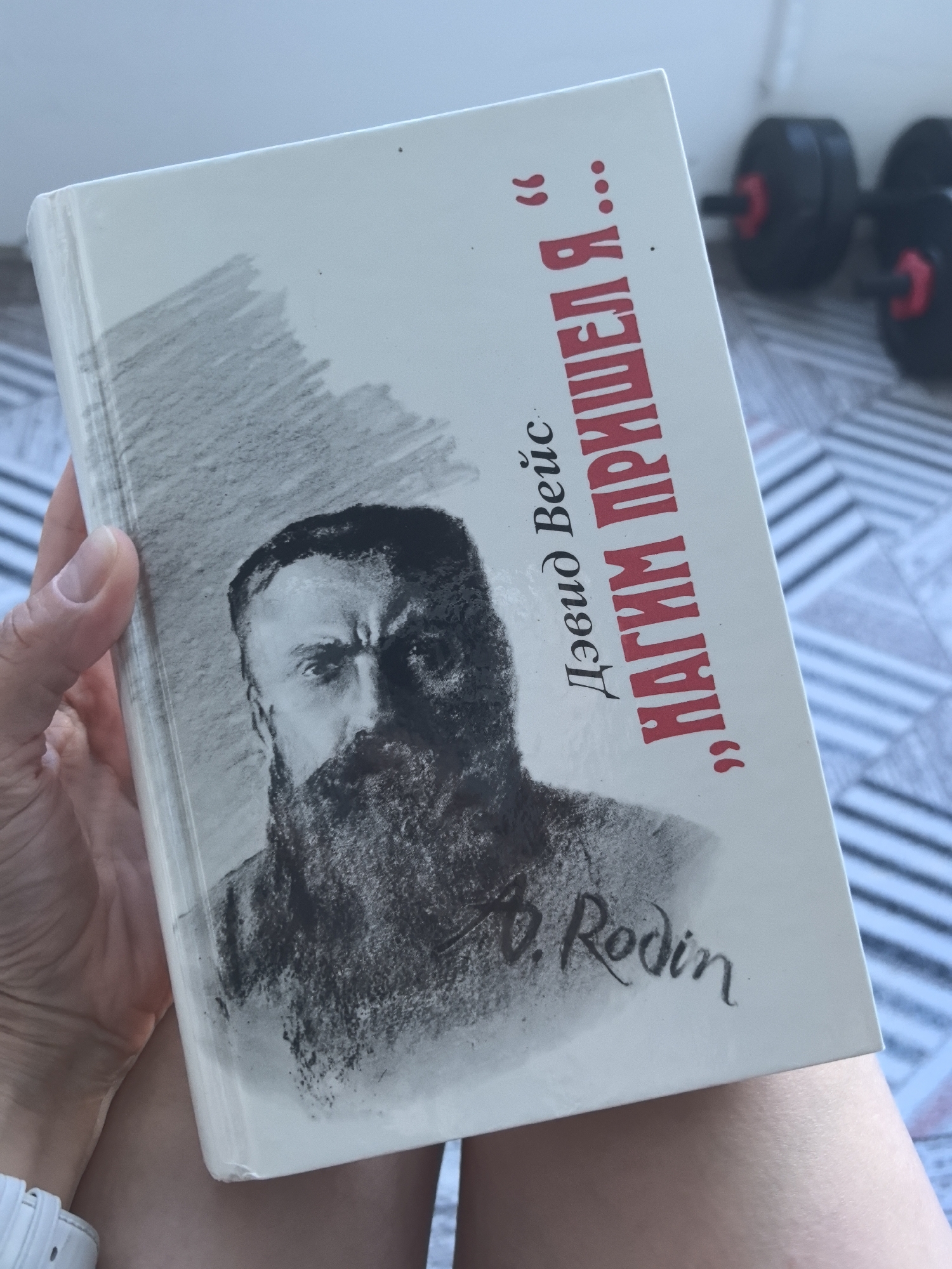 The image presents a close-up of a person's hand holding a book, likely indoors in Perm, Russia, given the language of the text. The book has a white cover featuring a charcoal-style illustration of a stern-looking, bearded man whose face is partly shadowed. Below the portrait, a cursive signature "B. Rodin" is visible, suggesting the subject is Auguste Rodin, the French sculptor.
The book's title, printed in prominent red Cyrillic letters, reads "„НАГИМ ПРИШЕЛ Я...”" (NAGIM PRISHEL YA...), which translates to "Naked I Came..." or "Naked I Arrived...". Above the title, the author's name is "Дэвид Вейс" (David Veis), or David Weiss, in black Cyrillic text.
The hand holding the book wears a white wristband. The book itself shows minor wear on its edges. In the blurred background, parts of black dumbbells with red accents are visible, implying a setting that might include a home gym or fitness equipment. A light-colored striped pattern, possibly from flooring or a mat, is also discernible in the background. The scene is illuminated by what appears to be natural daylight.