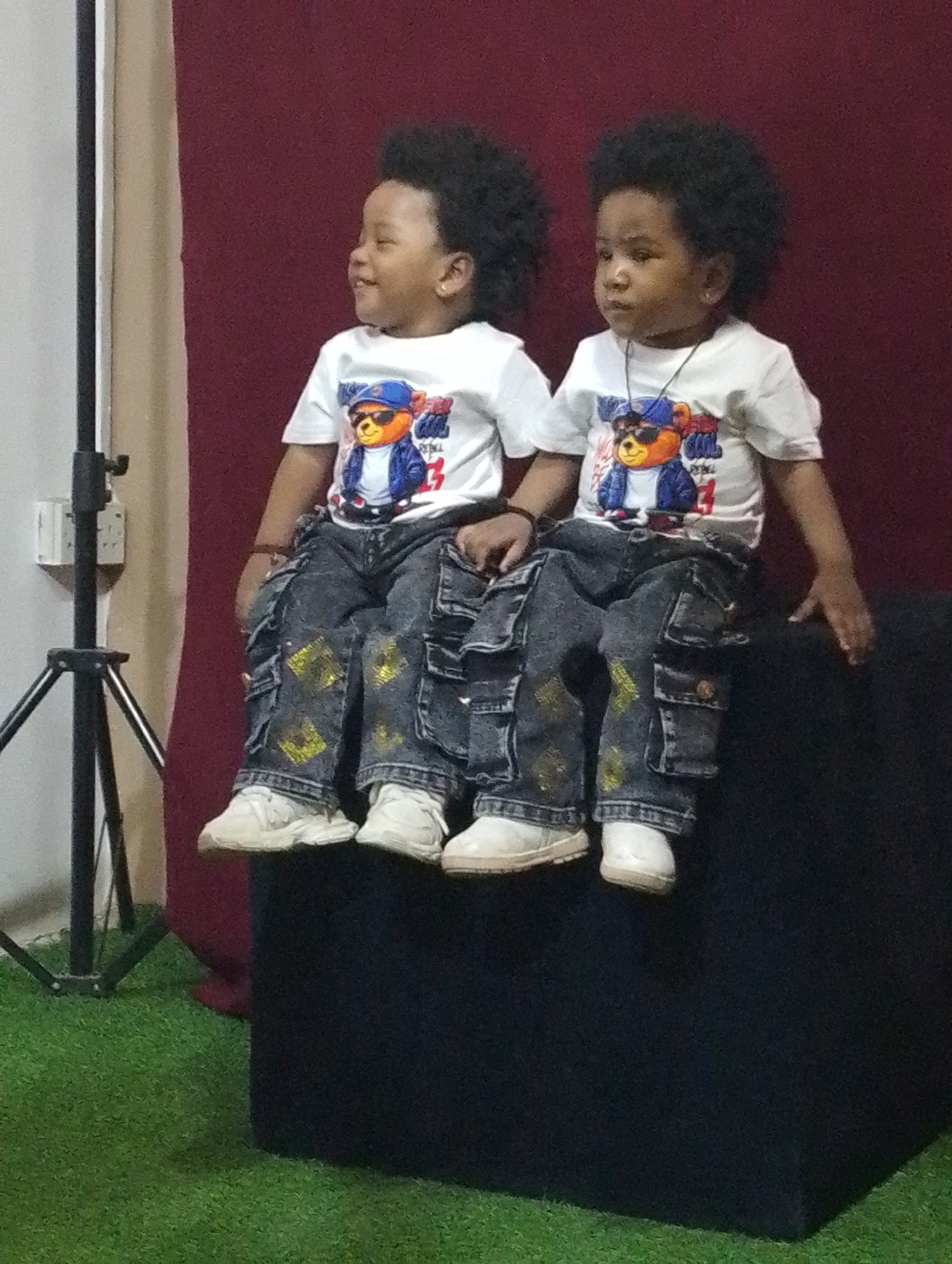 Two young children, appearing to be twins or siblings, are featured in an indoor portrait taken in Babban Giji, Nigeria. Both children have dark, curly hair and are dressed in matching outfits: white short-sleeved t-shirts adorned with a graphic of a bear wearing sunglasses, a blue jacket, and a blue cap. The word "REBEL" is visible on the t-shirt graphic, with "COOL" also partially discernible. They wear dark denim cargo pants with yellow or gold paint splatters and white sneakers.
The children are seated side-by-side on a dark, possibly black, rectangular block or stool. The child on the left is smiling and looking off to their left, while the child on the right has a more neutral expression, looking towards the camera.
The setting appears to be a photography studio or a designated photo area. Behind the children is a burgundy-colored fabric backdrop. To the left of the frame, a white wall or screen is visible, alongside a black tripod stand with an attached electrical outlet. The floor beneath them is covered with artificial green grass, adding a distinct element to the scene. The overall setup suggests a posed photo session.