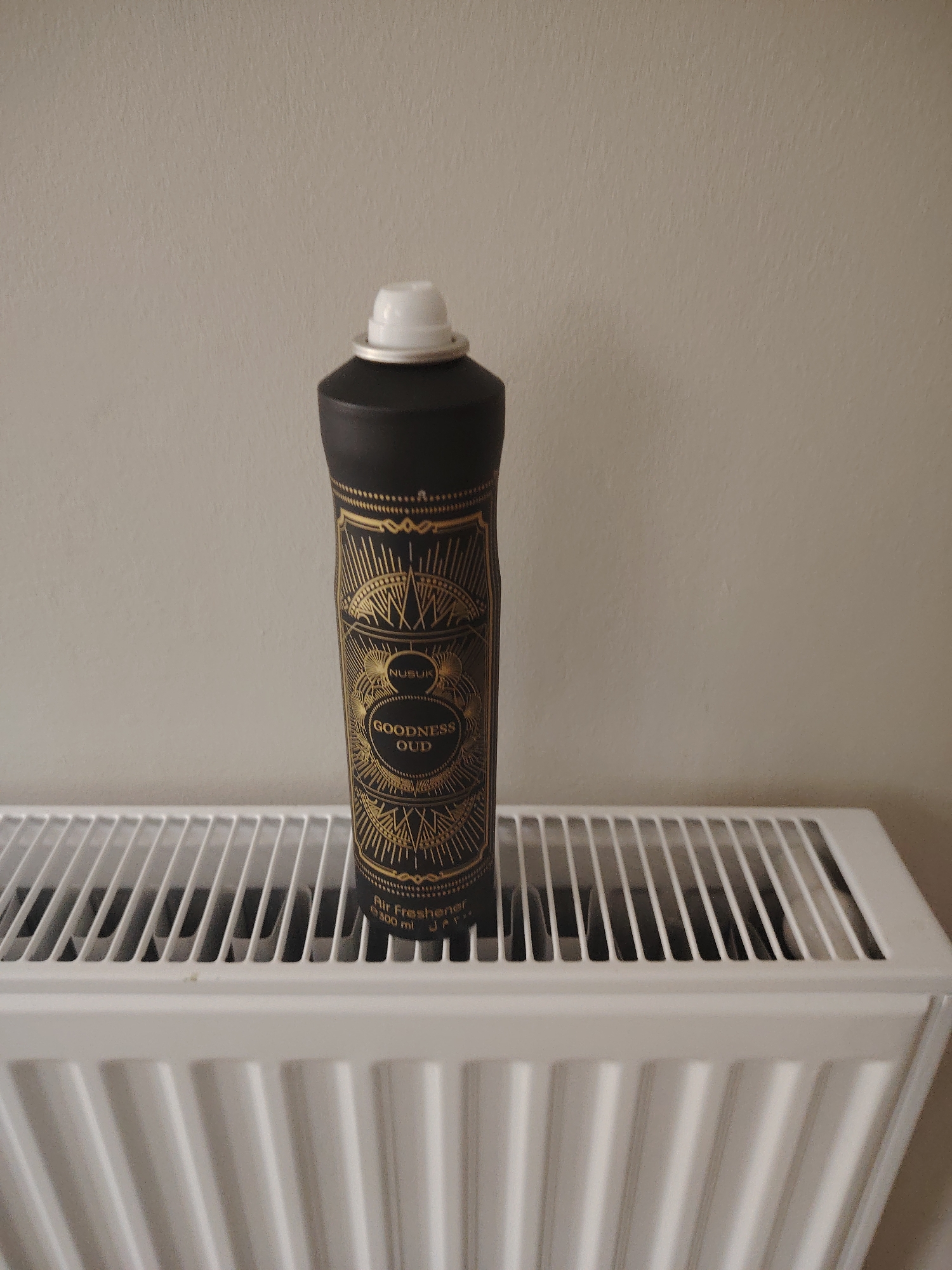 A black spray can, labeled "NUSUK GOODNESS OUD Air Freshener e 300 ml" with intricate gold art-deco style patterning, stands upright on the white slatted top grille of a modern panel radiator. The can features a white nozzle with a silver ring at its base. The radiator occupies the lower half of the frame, resting against a plain, light beige wall that fills the upper background. The scene is located in Huntingdonshire, United Kingdom.
