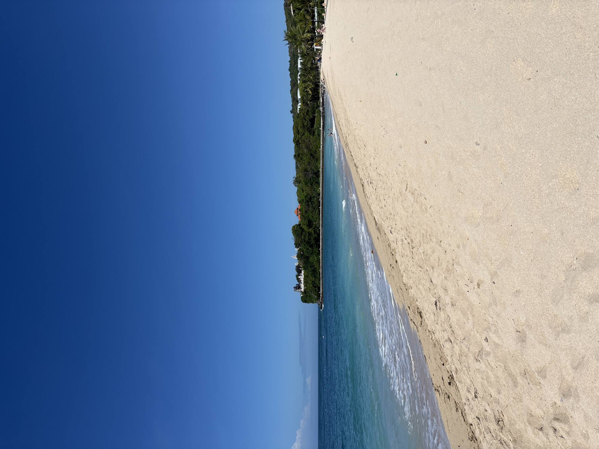 This is an eye-level, outdoor shot of a beach in Kuta Selatan, Indonesia. The photo depicts the sky on the left, and the beach and ocean on the right.
The sky is a uniform blue with no clouds. A narrow strip of land is visible, lined with trees and some buildings. The ocean gradually fades from a light blue near the shore to a darker blue further out. Small waves are visible. The sandy beach takes up the right half of the image. It's a light tan color with various footprints and debris scattered across the surface.
