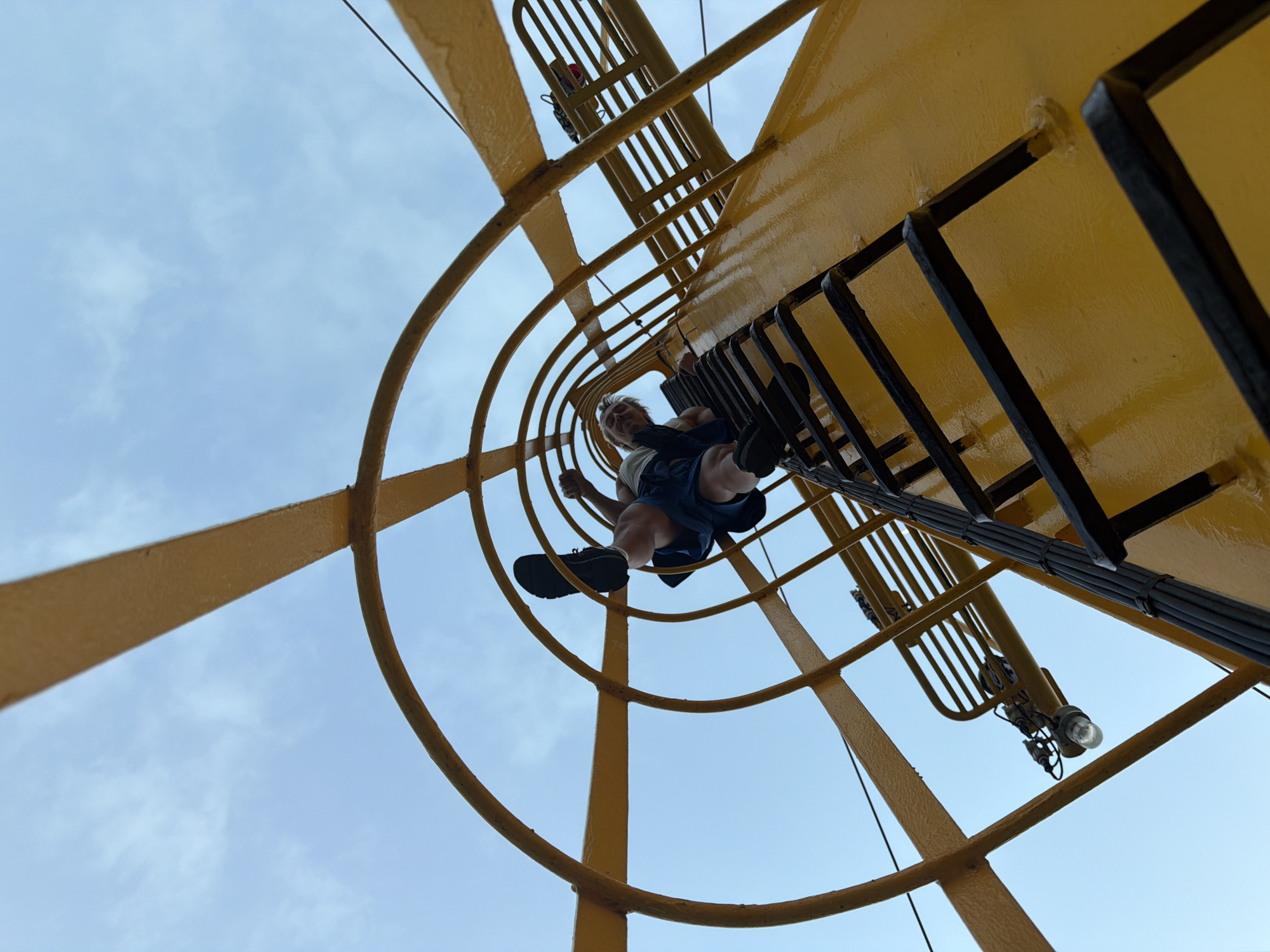 A person is climbing a yellow metal ladder on a clear day. The ladder has a cage-like structure around it, also made of yellow metal. The person is wearing blue shorts and a light-colored shirt, and is looking up at the camera. The background is a blue sky with some wispy clouds. The perspective is from below, looking up the ladder. There are some lights attached to the structure, and some black cables running along the side. The image appears to be taken in an outdoor industrial setting.