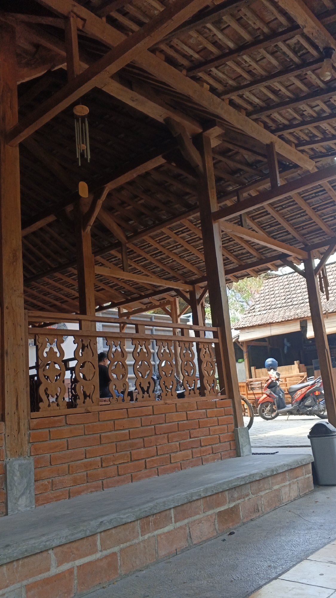 The image depicts an outdoor covered area, likely a restaurant or communal space, in Ngampel, Indonesia. The structure features a prominent wooden roof supported by beams and pillars. A decorative wooden railing, featuring intricate cut-out patterns, separates the covered area from an outdoor seating space. A brick wall forms the base of this railing. In the background, a tiled roof of another building is visible, along with a red motorcycle with a rider wearing a blue helmet. A grey trash can is positioned to the right of the frame. The ground in front of the structure is paved. A wind chime hangs from the wooden ceiling. The scene appears to be an ordinary day with no unusual activity.