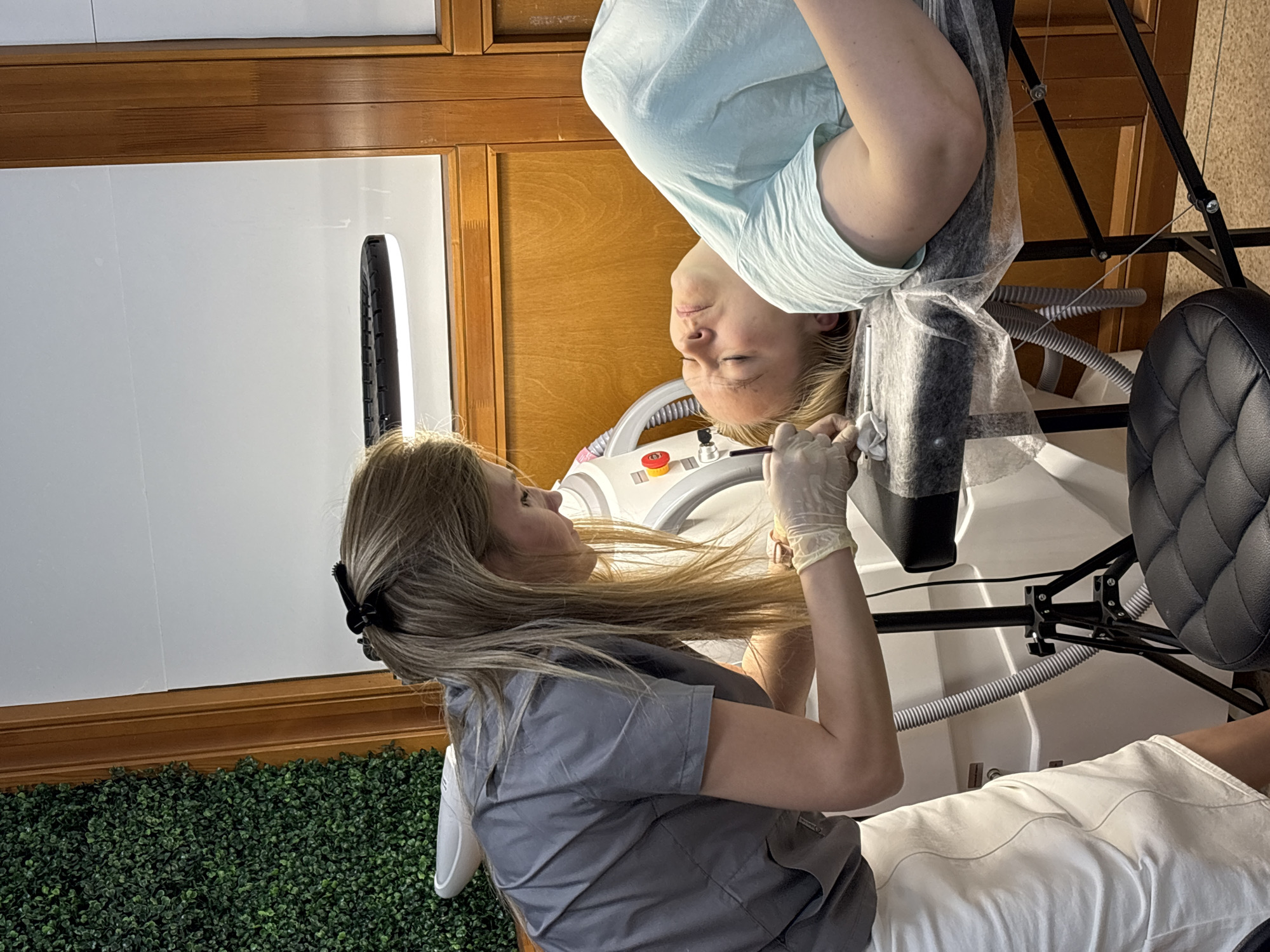 A woman in a gray scrub suit and latex gloves is providing a cosmetic treatment to a woman lying on a white examination table. The woman receiving treatment is wearing a light blue shirt. A piece of protective material covers part of the recipient’s face. The scene is illuminated by a ring light. A black, cushioned chair is present near the table. A green hedge-like wall decoration is behind the standing woman. The setting appears to be an aesthetician's office in an indoor setting. There is no evidence of it being in Chaikovsky, Russia.