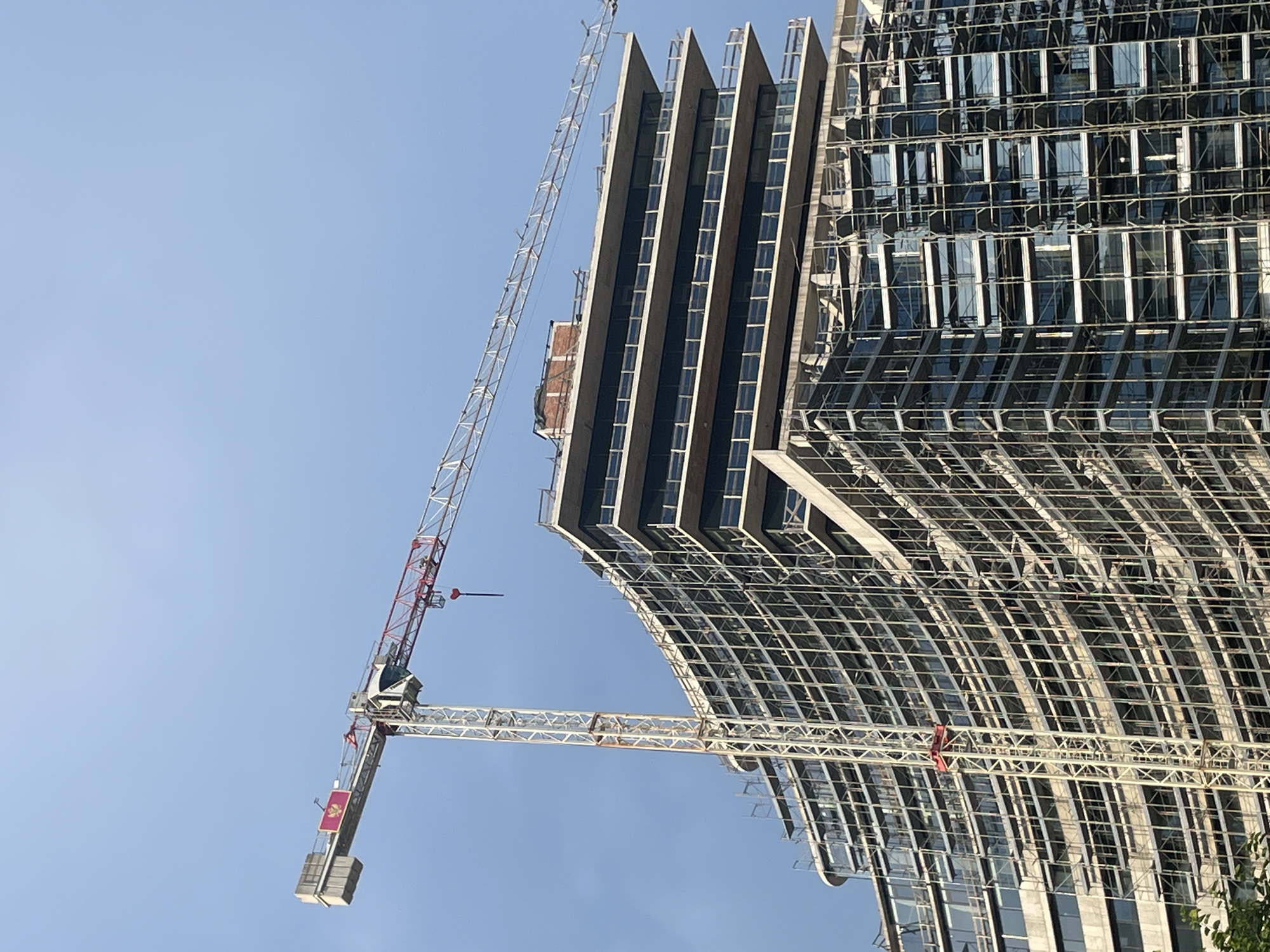 This is a low-angle, full-shot, eye-level, outdoor, daytime, vertical, close-up, medium-shot, medium-full-shot, full-length, aerial, full-body photograph of a construction crane and a modern building with a curved facade under construction. The building is clad in scaffolding and glass windows. The crane is positioned to the left of the building, with its jib extending over the top floors. The sky is clear and blue. The location is Budva, Montenegro.