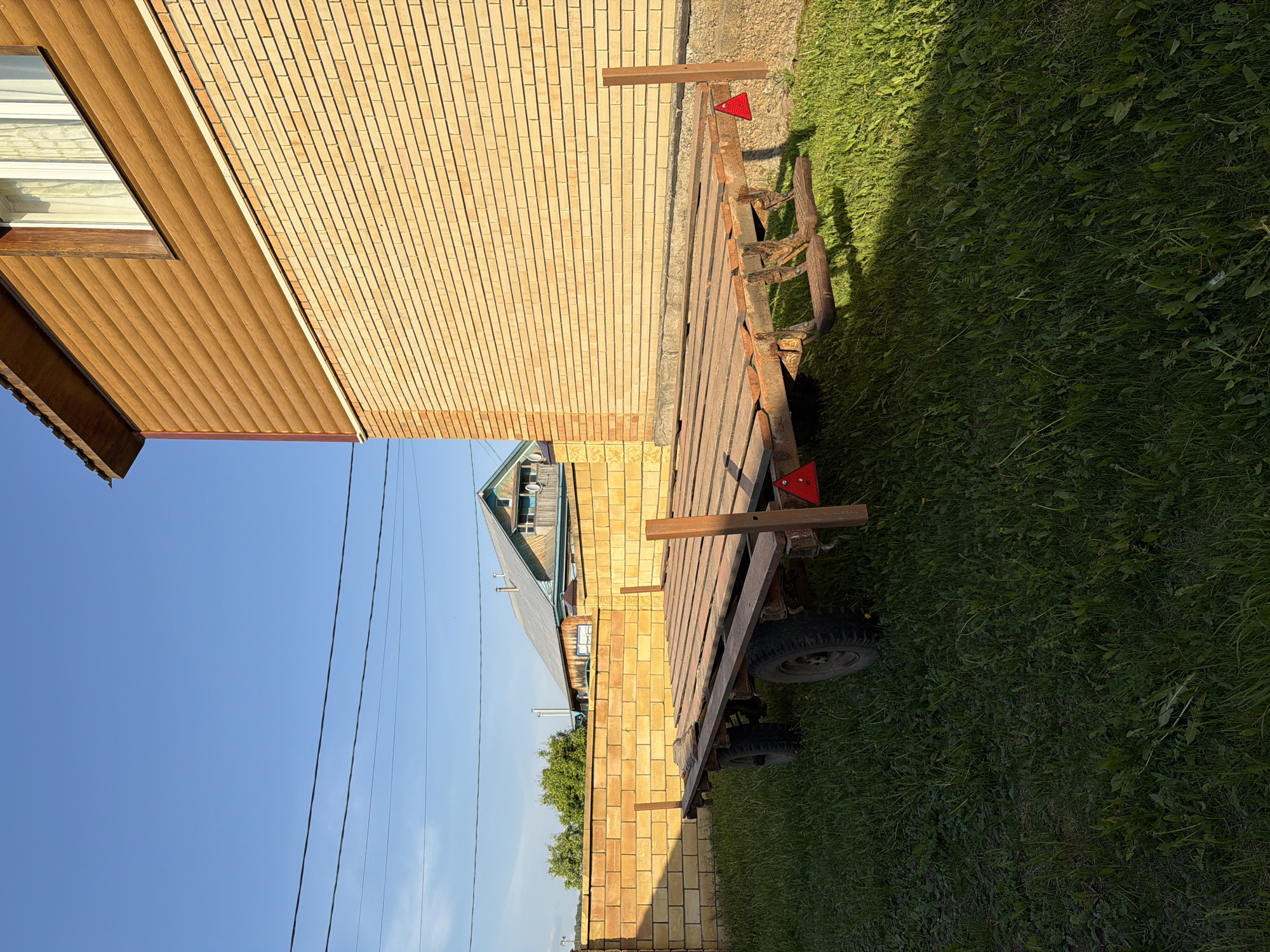 The scene depicts a trailer parked next to a building with brick and siding in what appears to be Bolsheustyikinskoye, Russia. The trailer has a wooden flatbed with a metal frame and appears to be positioned on grass beside the building. A section of blue sky is visible above, with power lines extending across the frame. The vegetation suggests a rural or suburban environment. No individuals are observable in this image.