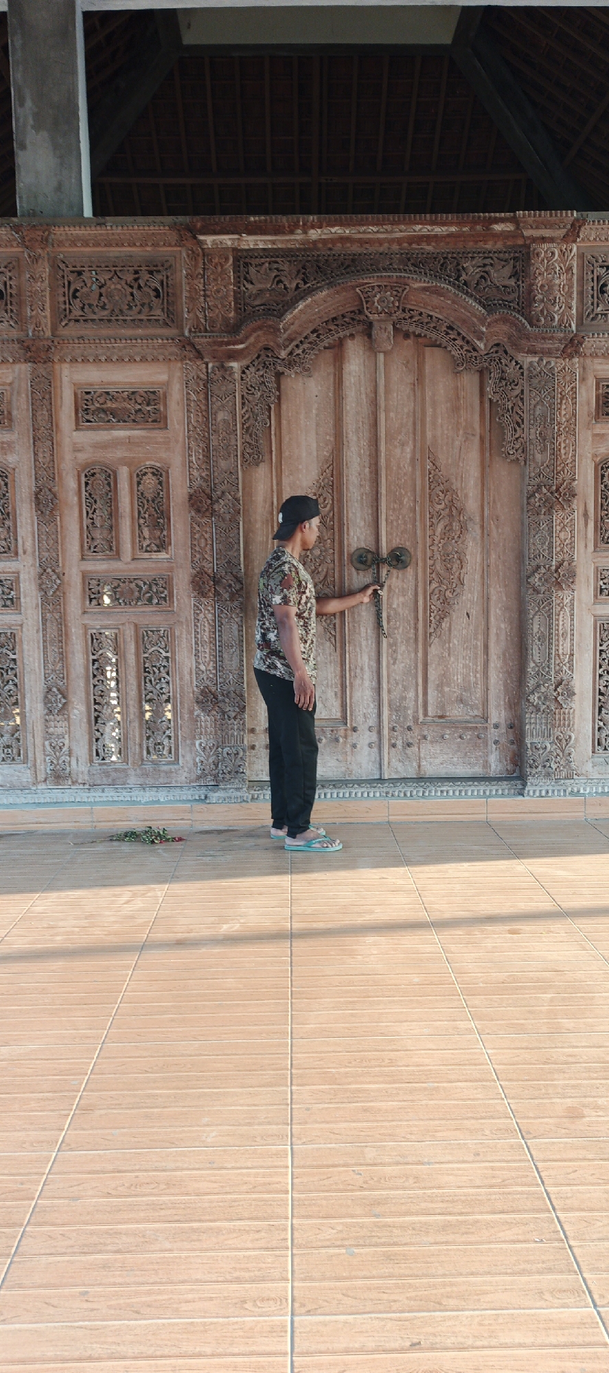 In the city of Jambearum, Indonesia, a young man stands in front of a large, intricately carved wooden door. He is wearing a camouflage t-shirt, black pants, a black baseball cap, and light blue flip-flops. His right hand is on the door handle, which is a circular metal object attached to a chain. The door is set into a wall that is also adorned with ornate carvings. The man is positioned in the center of the frame, facing the door, and appears to be either opening or closing it. In the foreground, a tiled floor reflects the warm sunlight. To the left of the man, a small pile of red flowers lies on the tiled ground. The background shows the interior of a structure with a wooden ceiling.