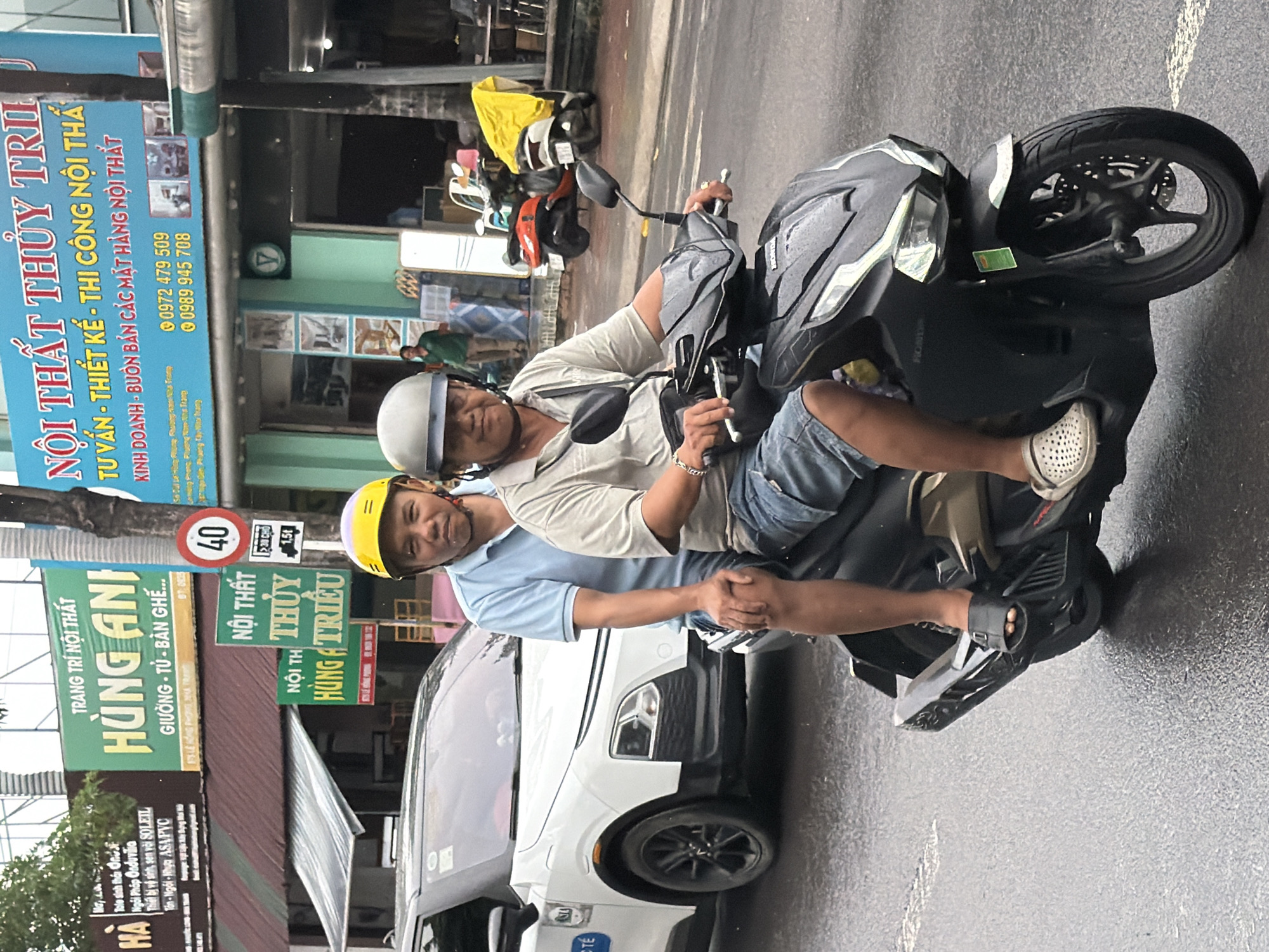 Two men are riding a black Honda scooter on a wet street in Nha Trang, Vietnam. The man in front is wearing a blue short-sleeved shirt and denim shorts, with a yellow helmet. The man behind him is wearing a grey short-sleeved shirt and denim shorts, with a grey helmet. Both are wearing sandals. The scooter has a green sticker on its front. In the background, there are several storefronts with signs in Vietnamese. One prominent sign is for "NỘI THẤT THỦY TRÌ," which translates to "Thuy Tri Furniture." Another sign says "TRANG TRÍ NỘI THẤT HÙNG ANH," meaning "Hung Anh Interior Decoration." A white car is parked on the left side of the street. A "40" speed limit sign is visible on a pole. The street appears to be an urban commercial area.