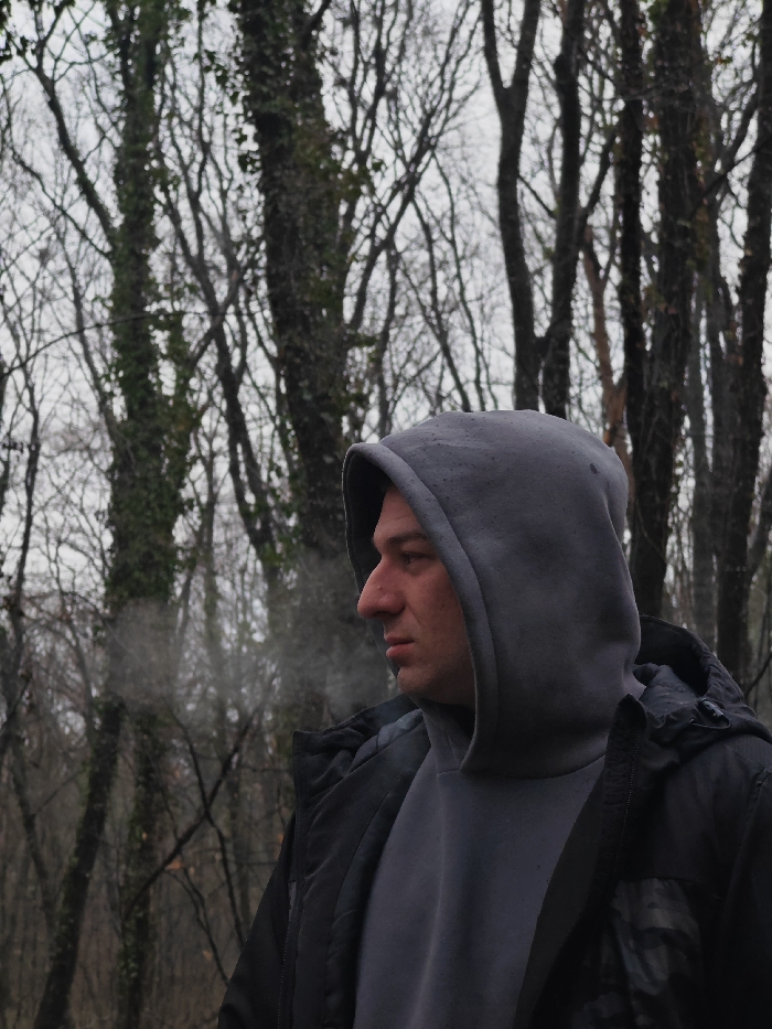 The image displays an adult male standing outdoors in a forest in Shumen, Bulgaria. The individual occupies the right side of the frame, visible from the chest up, and is shown in profile facing left. He wears a dark grey hooded sweatshirt with the hood pulled over his head, layered under a dark, likely black, padded jacket. Vapor or smoke is observed emanating from the area of his mouth, extending horizontally towards the left. His facial features include a prominent nose and a neutral expression. The background consists of a dense, leafless deciduous woodland, with tree trunks varying in texture and some covered in dark green ivy or moss. The sky is overcast and grey, indicating an ambient light condition. No other subjects or interactions are present in the scene.