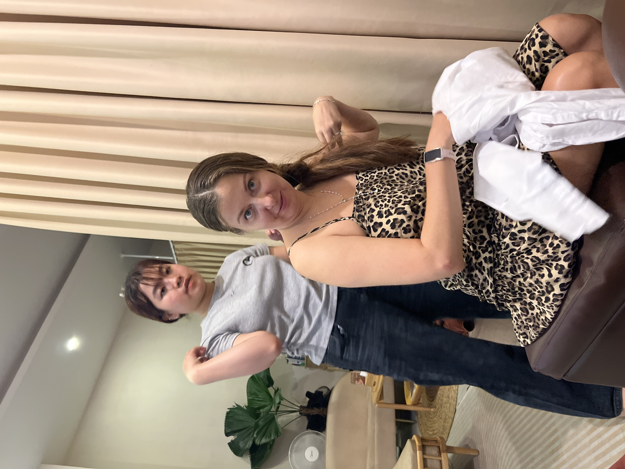 Two women are captured in an indoor setting. One woman is seated on what appears to be a dark brown leather couch, wearing a leopard-print dress and a smart watch. She is holding a white object in her left hand. The other woman, wearing a gray shirt and dark jeans, stands behind and slightly to the side of the seated woman. In the background are neutral-toned curtains, a potted plant, a floor fan, and various pieces of furniture. The location is not specified.