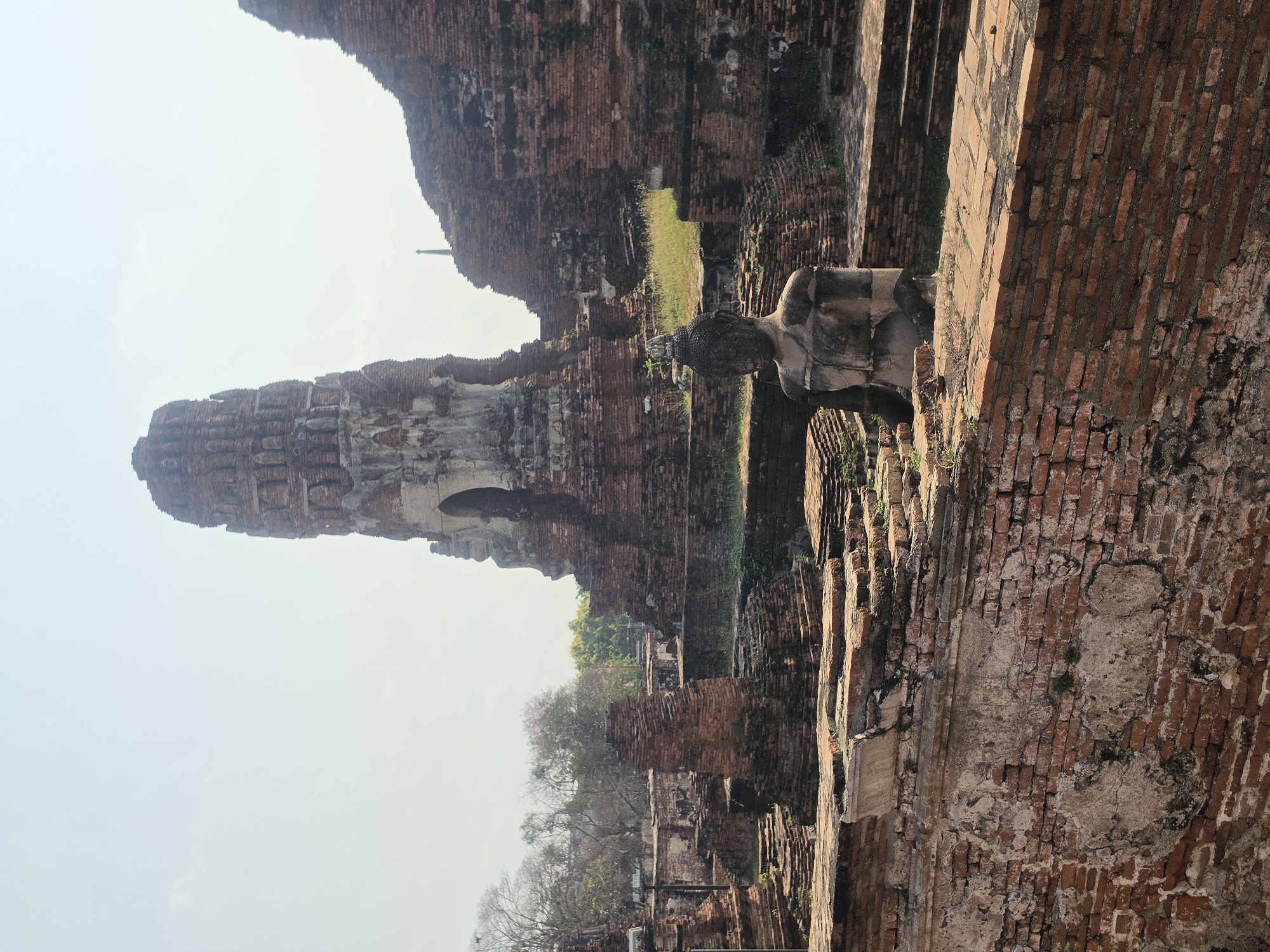 The image shows the Wat Yai Chaimongkol temple ruins in Phra Nakhon Si Ayutthaya City Municipality, Thailand. The ruins are made of red brick. There is a tall tower-like structure. In the foreground, there is a headless Buddha statue. There is grass and trees in the background. The sky is clear.