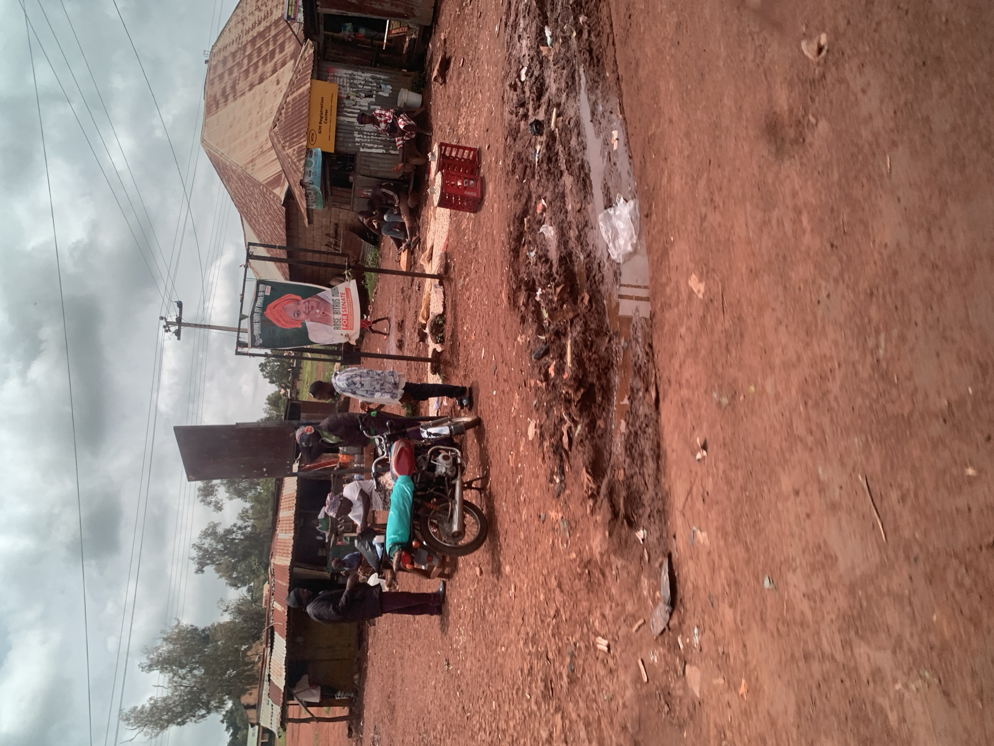 The image depicts an outdoor scene in Hoss, Nigeria, characterized by a dusty, reddish-brown dirt road with puddles of muddy water. In the background, there are several corrugated metal structures that appear to be shops or market stalls, some with tin roofs. Power lines are visible overhead.
In the mid-ground, a group of people are gathered. A man in a dark jacket and cap stands near a parked motorcycle, which is covered with a green tarp. To his right, a woman wearing a white headscarf and white clothing is seated, and another person is partially visible behind her. Further to the right, a man in a light-colored patterned shirt stands. There's a large political poster on an easel displaying a woman wearing a red headwrap and the text "ROSE BITRUS MU... FOR SENATE." Next to this poster is a yellow sign that reads "MTN SIM Registration Center." Two red plastic crates are placed on the ground near the registration center.
In the background, more people are seated and standing around the market stalls. Some of the structures appear weathered and in disrepair. Trees with sparse foliage are visible behind the buildings. The overall impression is a bustling, albeit somewhat rudimentary, commercial area.
