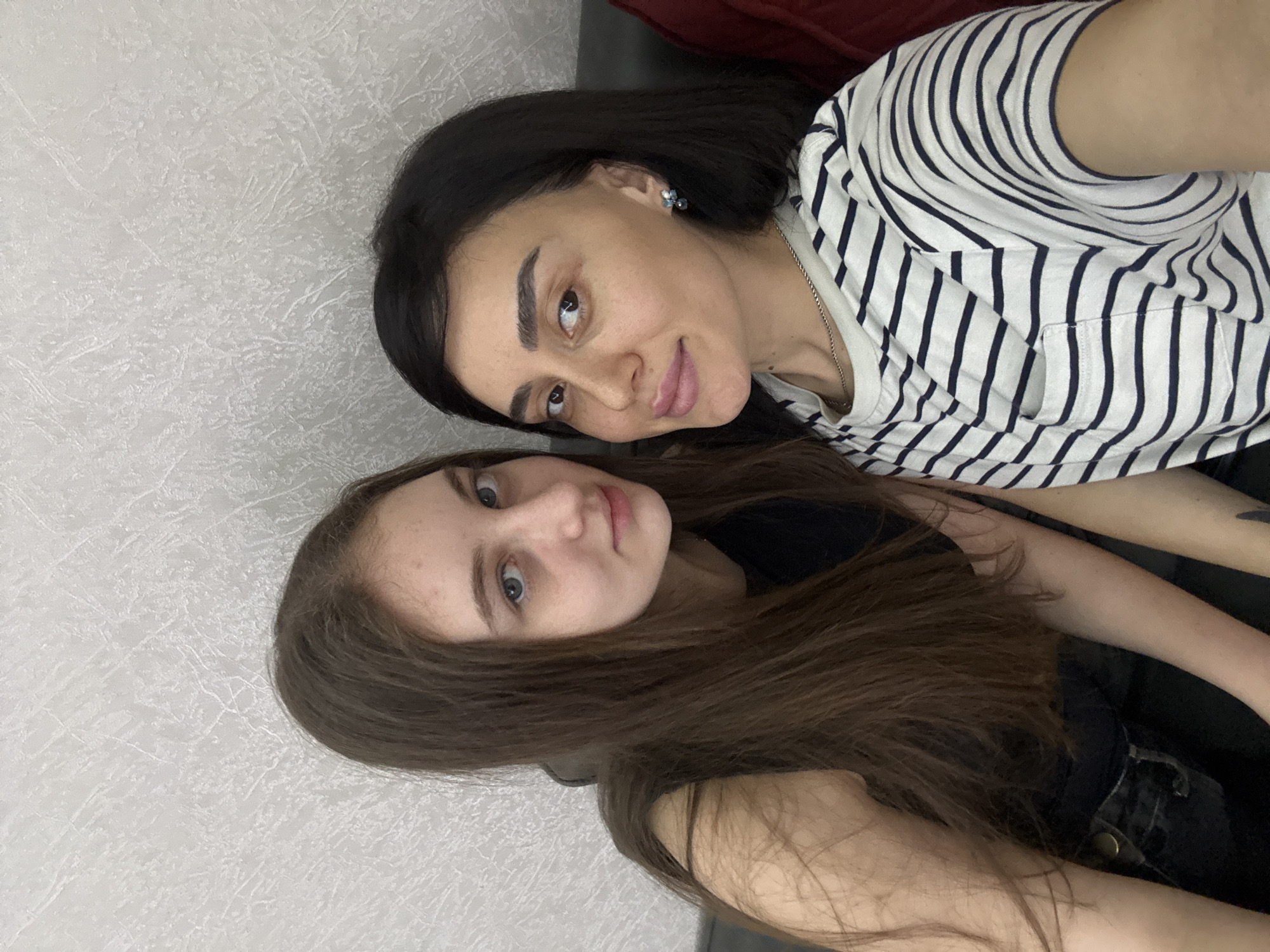 Two females are visible, lying next to each other. The woman on top has black hair and is wearing a white and black striped shirt. The woman on the bottom has long brown hair and is wearing a black top. The background consists of a textured light-colored wall and a dark red surface. The location is indoors. The city and country (Tbilisi, Georgia) are not evident from the image.