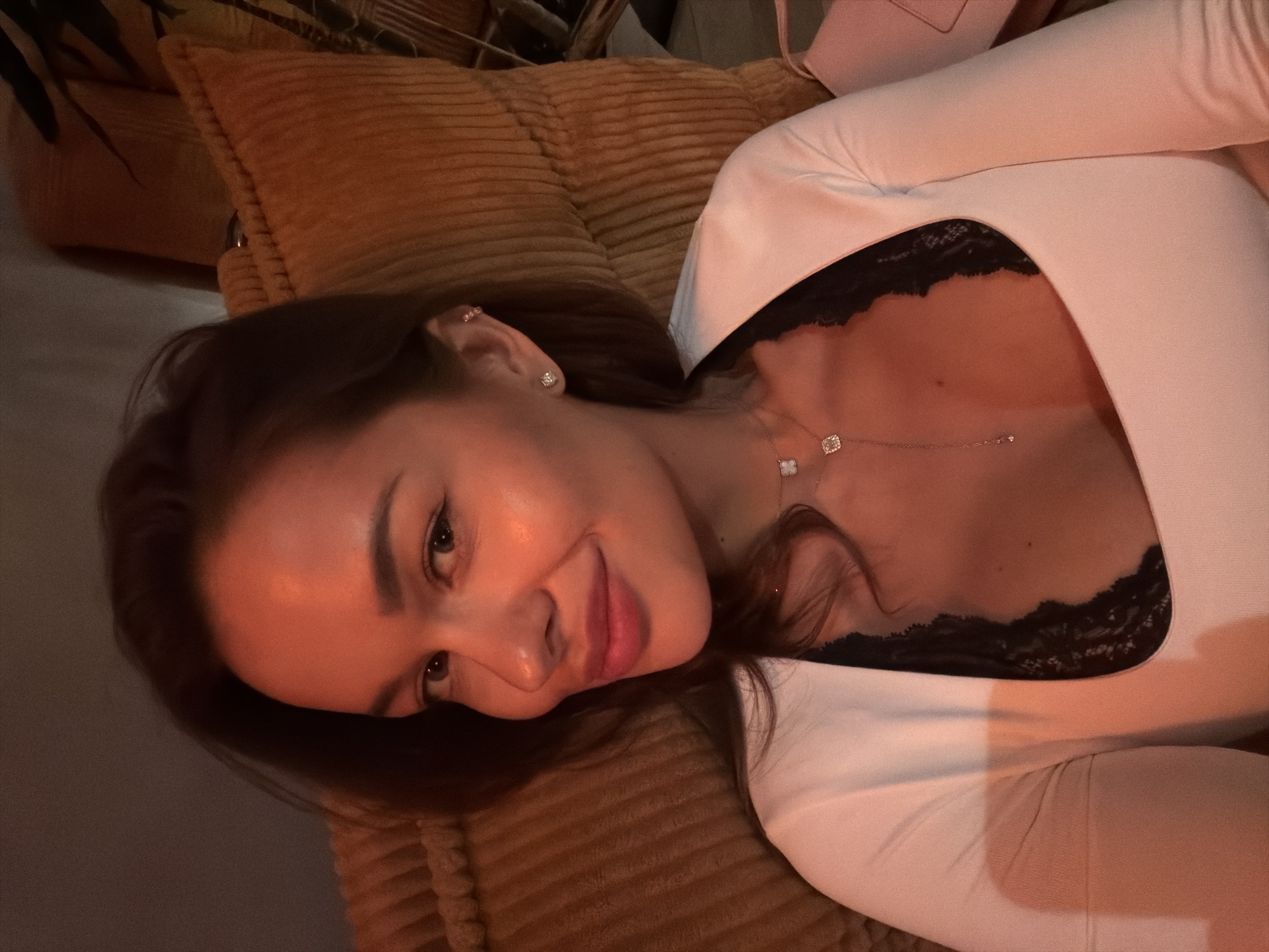 This close-up, angled portrait features a young woman lying down in an indoor setting. Her head is positioned towards the left side of the frame, tilted, with her body extending diagonally. She has dark hair, dark eyes, and a soft, pleasant smile directed towards the camera. Warm, ambient lighting casts an orange glow across her face and upper chest, suggesting an evening or nighttime atmosphere.
The woman wears small, sparkling stud earrings, with one visible on her left ear. A delicate necklace with two distinct pendants – one square or clover-shaped, and another smaller, elongated one – adorns her décolletage. She is dressed in a light-colored, long-sleeved top, possibly white or pale pink, beneath which a black lace bra or camisole is visible.
Behind her head and partially under her body is a textured, corduroy-like pillow or cushion in a warm orange-brown color. Another similar textured surface is visible further back. The overall scene conveys a cozy and relaxed indoor environment, potentially a living room or bedroom. The woman appears relaxed and content, likely taking a selfie. The location context for this media file is Moscow, Russia. There is no visible text in the image.