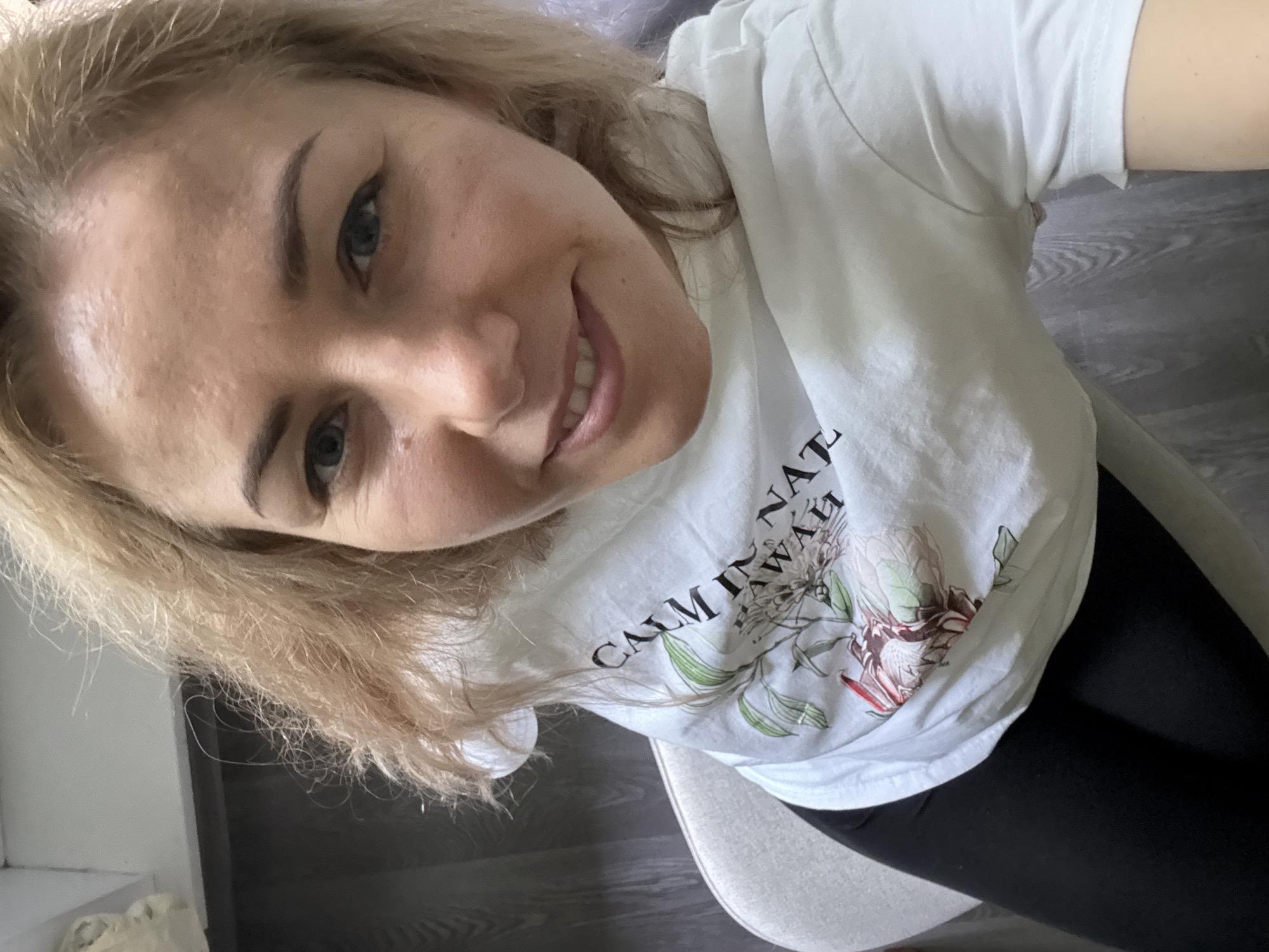 A medium shot depicts a blonde woman from a high-angle perspective, seemingly taken as a selfie. She is wearing a white t-shirt with a graphic design incorporating flowers and text that reads "CALM IN HAWAII." Below her shirt, black leggings are visible. The woman has fair skin, light-colored eyes, and is smiling. She is sitting on a chair with a neutral-colored seat. The background includes a wooden floor and a corner of a white wall, but no specific location in Moscow, Russia, can be determined from these limited environmental details.