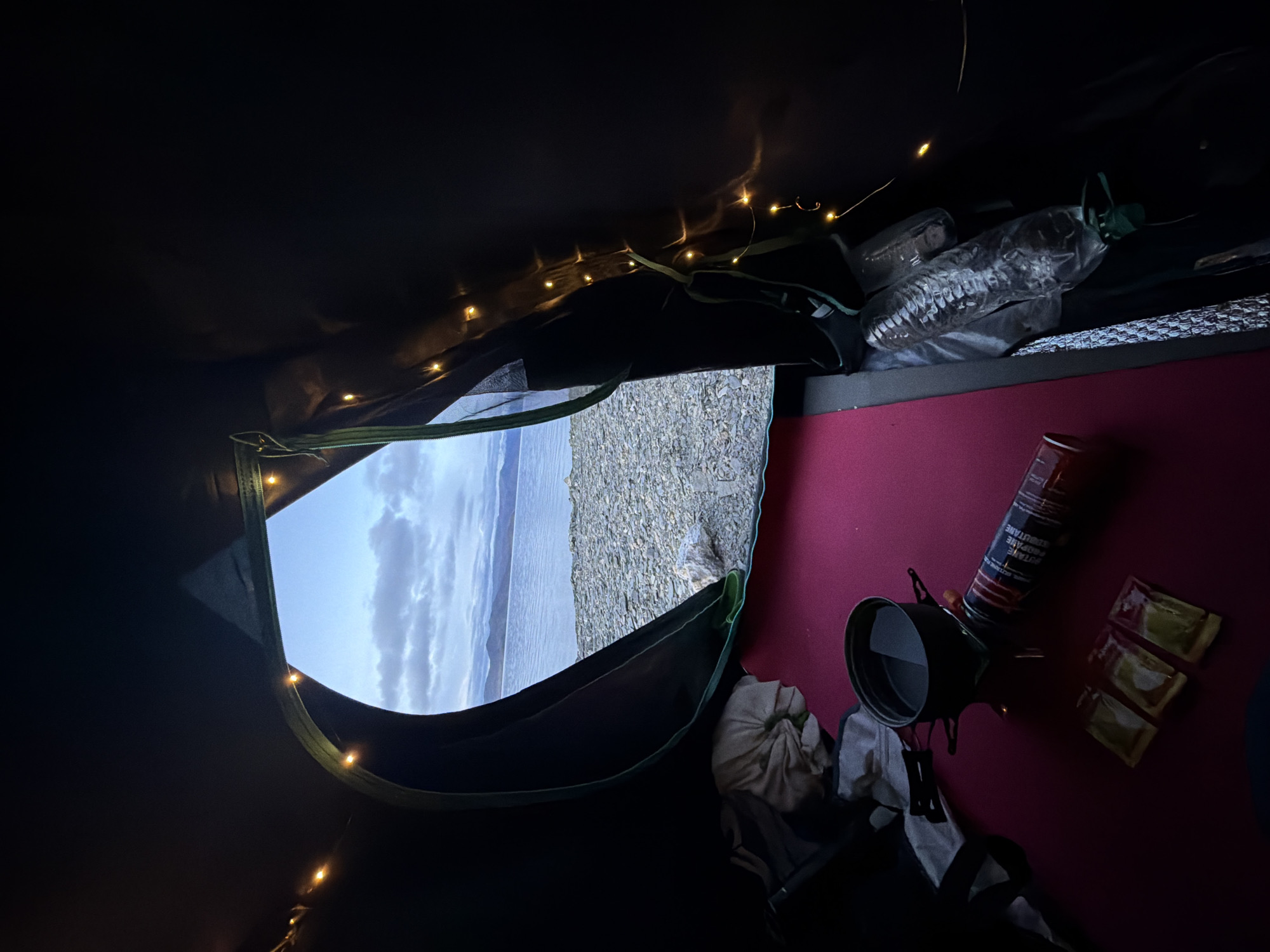 The image shows the interior view of a tent facing outwards. The tent opening reveals a body of water with a rocky shore in the foreground and mountains in the distance. The sky is cloudy and appears to be twilight. Inside the tent, a pink sleeping mat is laid out. On the mat, there is a small camping stove with a pot of water on it, a fuel canister, and three small packets of what appear to be food or seasoning. To the left, there is a crumpled piece of fabric. Along the right side of the tent opening, a string of small yellow fairy lights is visible. The overall impression is of a simple camping setup in a scenic, remote location. The location is described as being in Unknown, India.