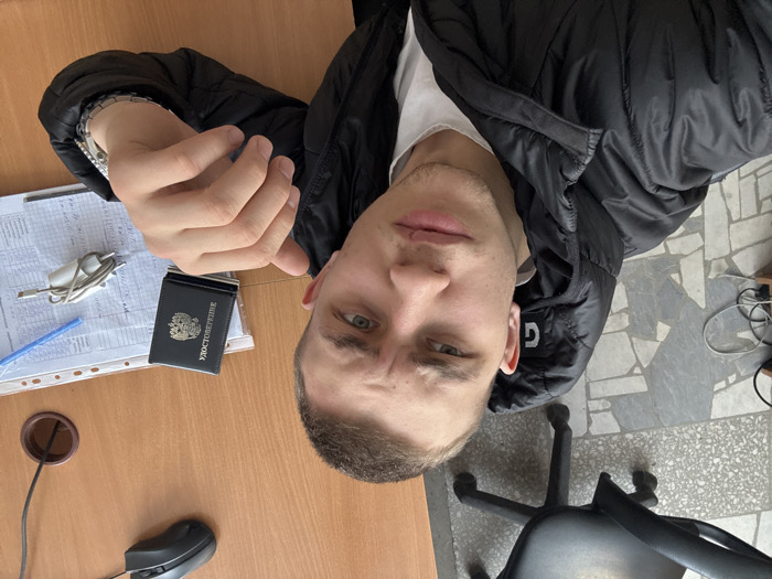 The image depicts a male individual indoors in Chaikovsky, Russia, captured in a close-up, selfie-style perspective. The subject, a young adult with short dark hair and light eyes, wears a black puffer jacket over a white garment. A minor scar or mark is visible on his upper lip. He has a neutral to slightly contemplative facial expression, with subtly furrowed brows. A silver-toned wristwatch is present on his left wrist, and his left hand is raised with fingers slightly curled.
The individual is positioned at a wooden-textured desk. On the desk surface, to the right of the subject, are a black computer mouse, a dark-colored identification booklet labeled "УДОСТОВЕРЕНИЕ" featuring the Russian Federation coat of arms, a stack of white papers, a blue pen, and a coiled white USB charging cable with an attached power adapter. A circular cable management grommet is visible in the desk surface.
The background, particularly to the left of the subject, reveals a partially visible black office chair with a five-star wheeled base. The floor is composed of light-toned mosaic or terrazzo tiles. Electrical wiring is visible on the floor near the chair. The environment indicates an office or administrative setting.