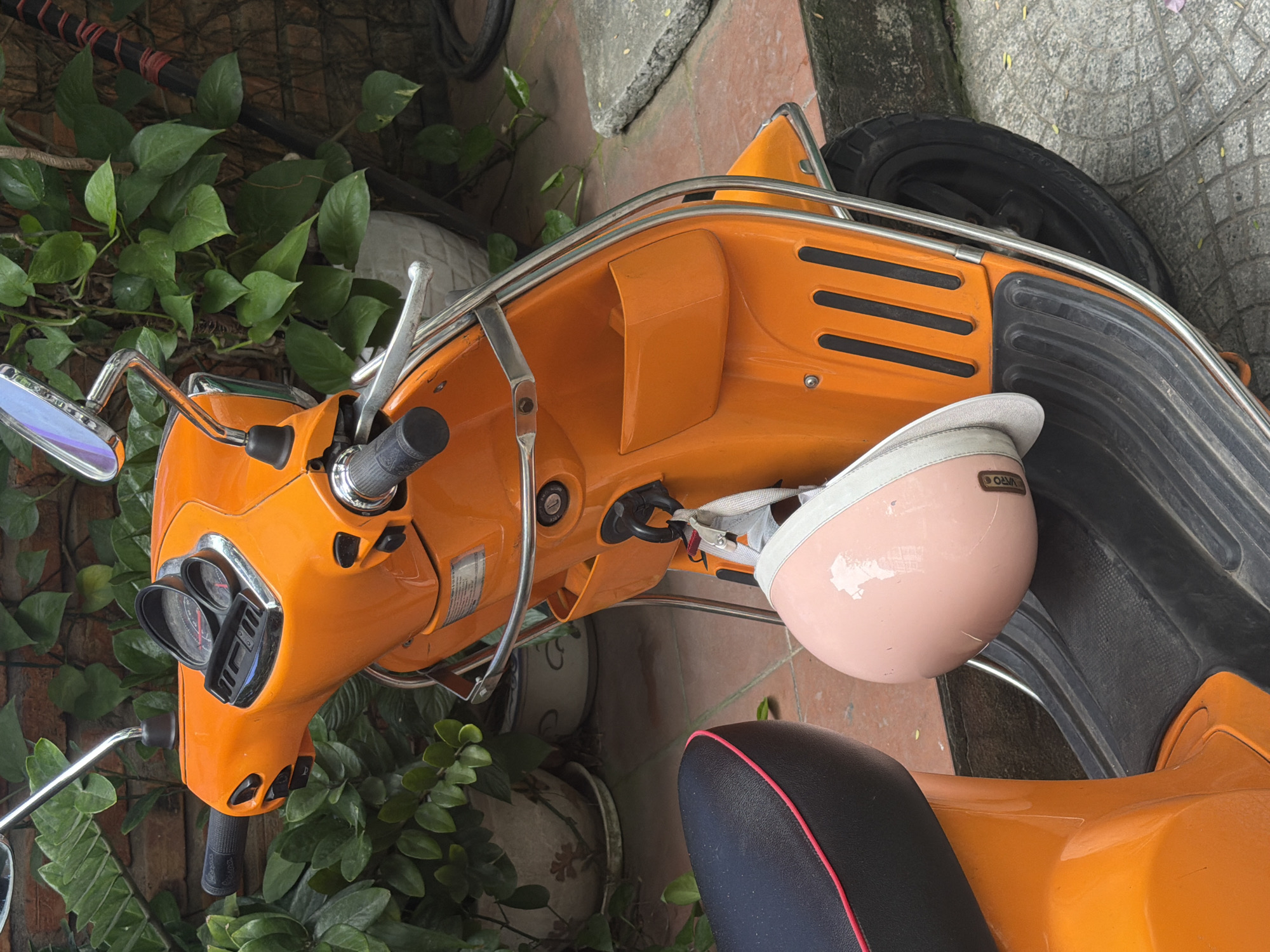 The image shows a parked, bright orange Vespa scooter on a tiled surface with green foliage visible nearby. A pink helmet rests on the scooter's seat. The location is suggested to be in Đà Nẵng, Vietnam, based on the urban setting and Vietnamese signage.