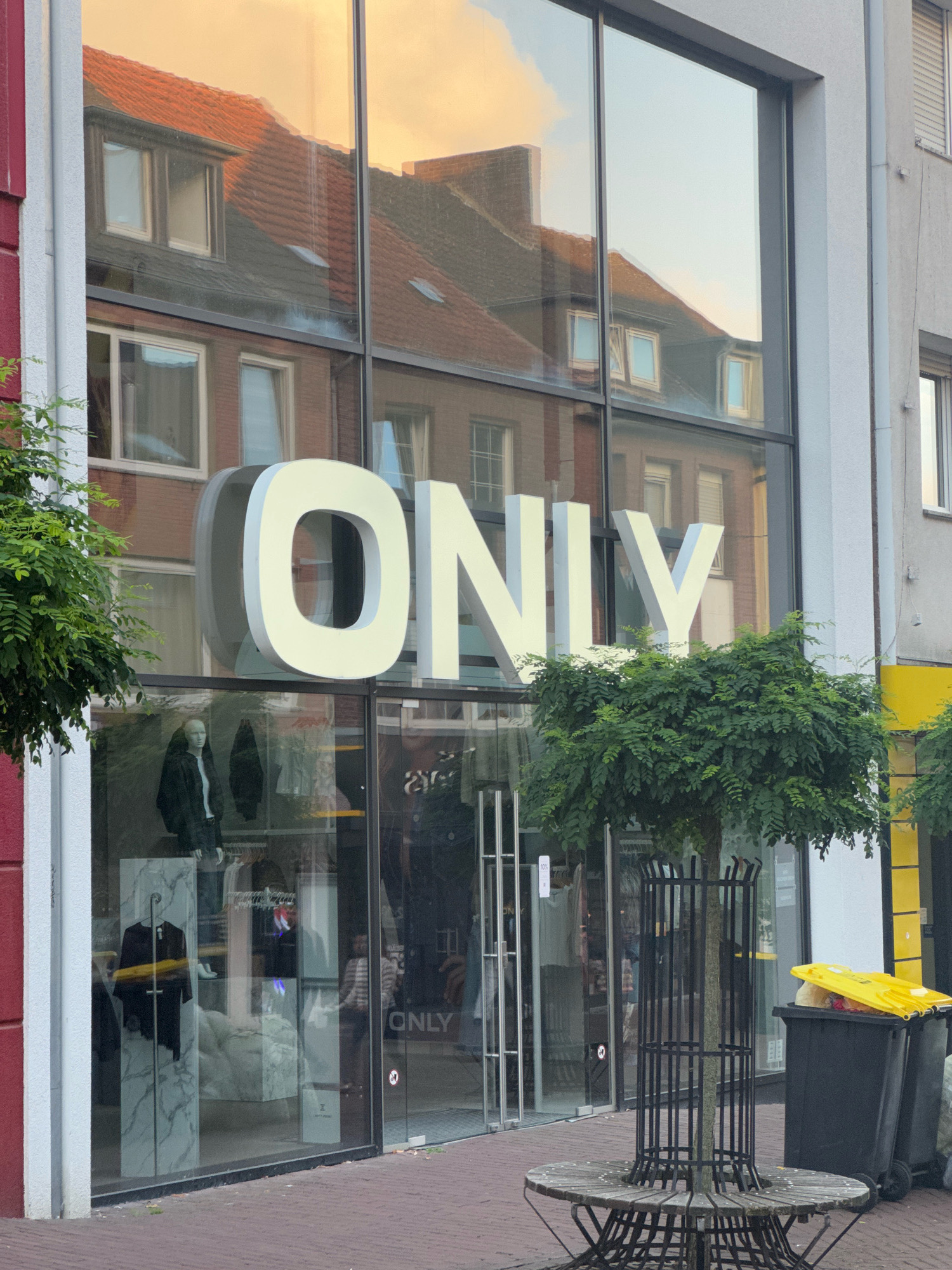 The image shows the storefront of an "ONLY" clothing store in Cleves, Germany. The store has a large glass facade with the brand name "ONLY" displayed in large, white, three-dimensional letters above the entrance. Reflections of buildings and the sky are visible on the glass. In the foreground, there is a tree with a protective metal grate and a circular wooden bench around its base. To the right of the tree, a black trash bin with a yellow lid is visible. The store window displays mannequins dressed in clothing, as well as racks of clothes. The street in front of the store is paved with red brick. The overall scene depicts a commercial street in a European city.