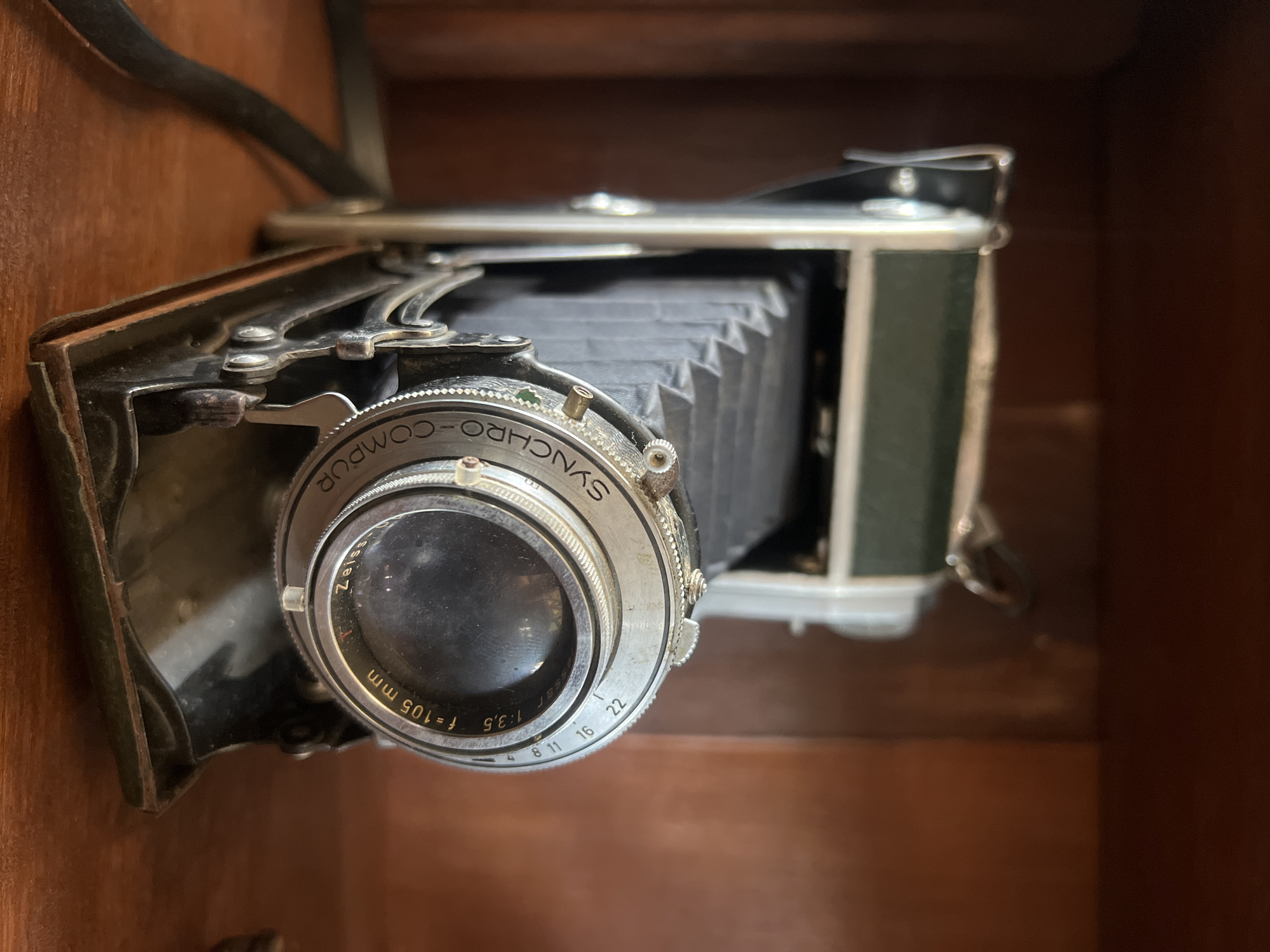 An old, green and silver bellows camera with a dark bellows is positioned on a wooden surface. The camera has a circular lens with markings that include "SYNCHRO-COMPUR," "f=105 mm," and aperture settings like "1.3.5," "8," "11," "16," and "22." The camera is on a wooden shelf or table. The background is a dark wooden surface. The image is a close-up shot of the camera, with the focus on the lens and bellows. No people are present in the image. The camera appears to be in good condition, despite its age. The lighting is natural, with soft shadows indicating an indoor setting. There is no information to suggest the location is Ubud District, Indonesia.