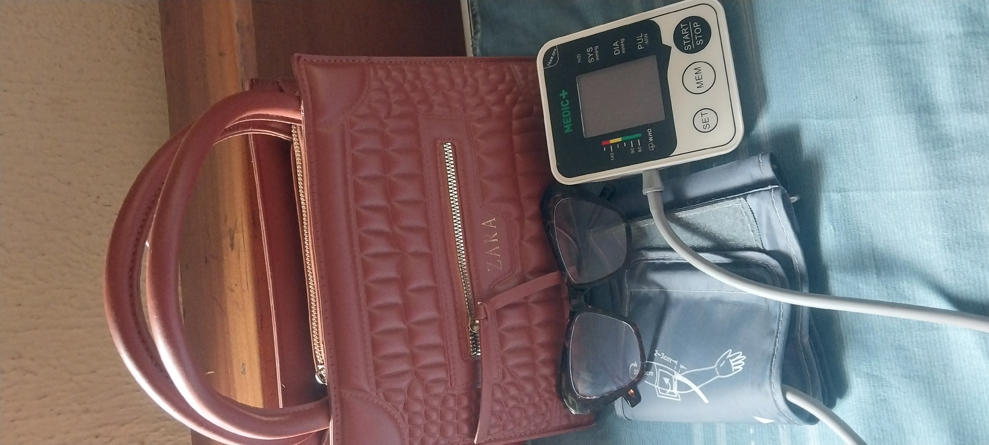 This media file shows a collection of personal items laid out on a textured blue fabric surface, possibly a bedspread or a tablecloth. The objects include a maroon textured handbag with "ZARA" embossed on it, a pair of dark-rimmed glasses, and a digital blood pressure monitor with an attached arm cuff. The blood pressure monitor is white with black and green accents, displaying readings for SYS (systolic), DIA (diastolic), and PUL (pulse). It also features buttons for "START/STOP", "MEM" (memory), and "SET", as well as a WHO classification chart and the brand name "MEDIC+". The arm cuff is gray and shows a diagram indicating proper placement around the upper arm. The scene is indoors, with a wooden surface visible to the left, possibly a desk or shelf. The lighting suggests it could be daytime. There are no people visible in the frame. The overall impression is one of items belonging to an individual, perhaps gathered before or after use. The location context provided is Onitsha, Nigeria.
