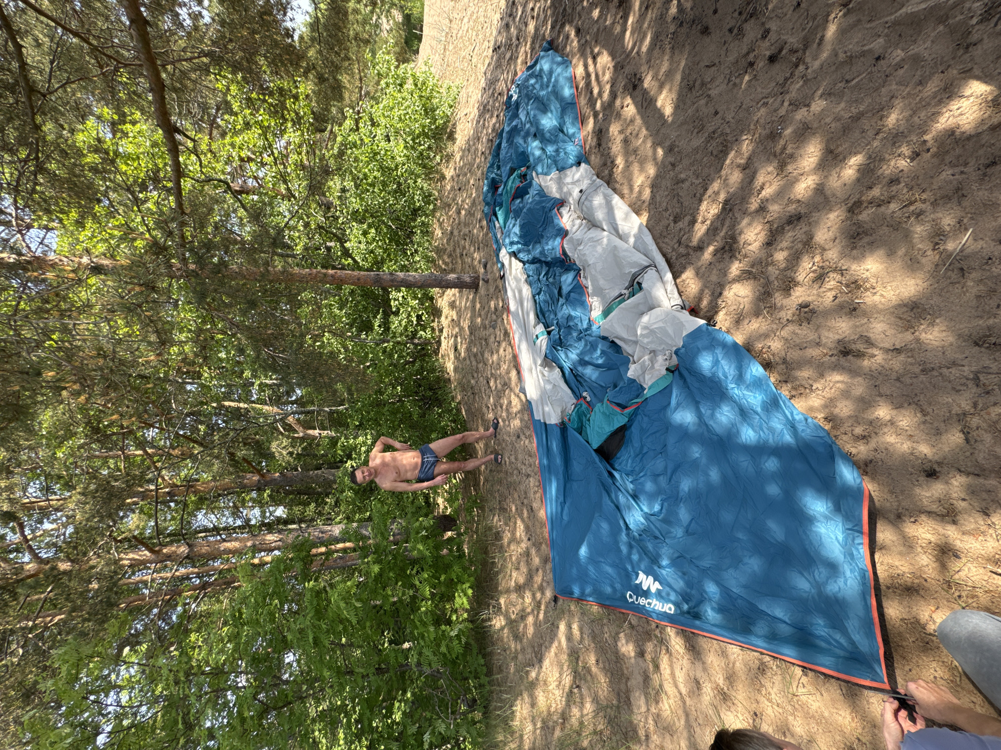 This media file depicts an outdoor scene in a natural, forested setting, consistent with Zelenaya Roshcha, Russia. The time of day appears to be sunny midday or early afternoon, as evidenced by the bright light and distinct, long shadows cast by trees across the sandy ground.
The primary subjects are a man and a camping tent. On the left side of the frame, a shirtless man, wearing blue swim shorts and dark sandals, stands casually amidst green foliage and tall pine trees. He has dark hair and appears to be looking towards the camera, with a relaxed posture.
To the right, a large, blue and white camping tent is partially laid out on the sandy ground, suggesting it is either being set up or packed away. The visible text "Quechua" with its logo is clearly printed on the tent fabric. In the bottom right corner, the lower legs and feet of another person are visible, indicating they are actively engaged with the tent, possibly securing or folding it.
The setting is characterized by sandy soil underfoot, interspersed with patches of green leafy bushes and numerous tall pine trees that form a dense canopy overhead. The overall atmosphere suggests an outdoor recreational activity like camping or a picnic in a natural, wooded area.