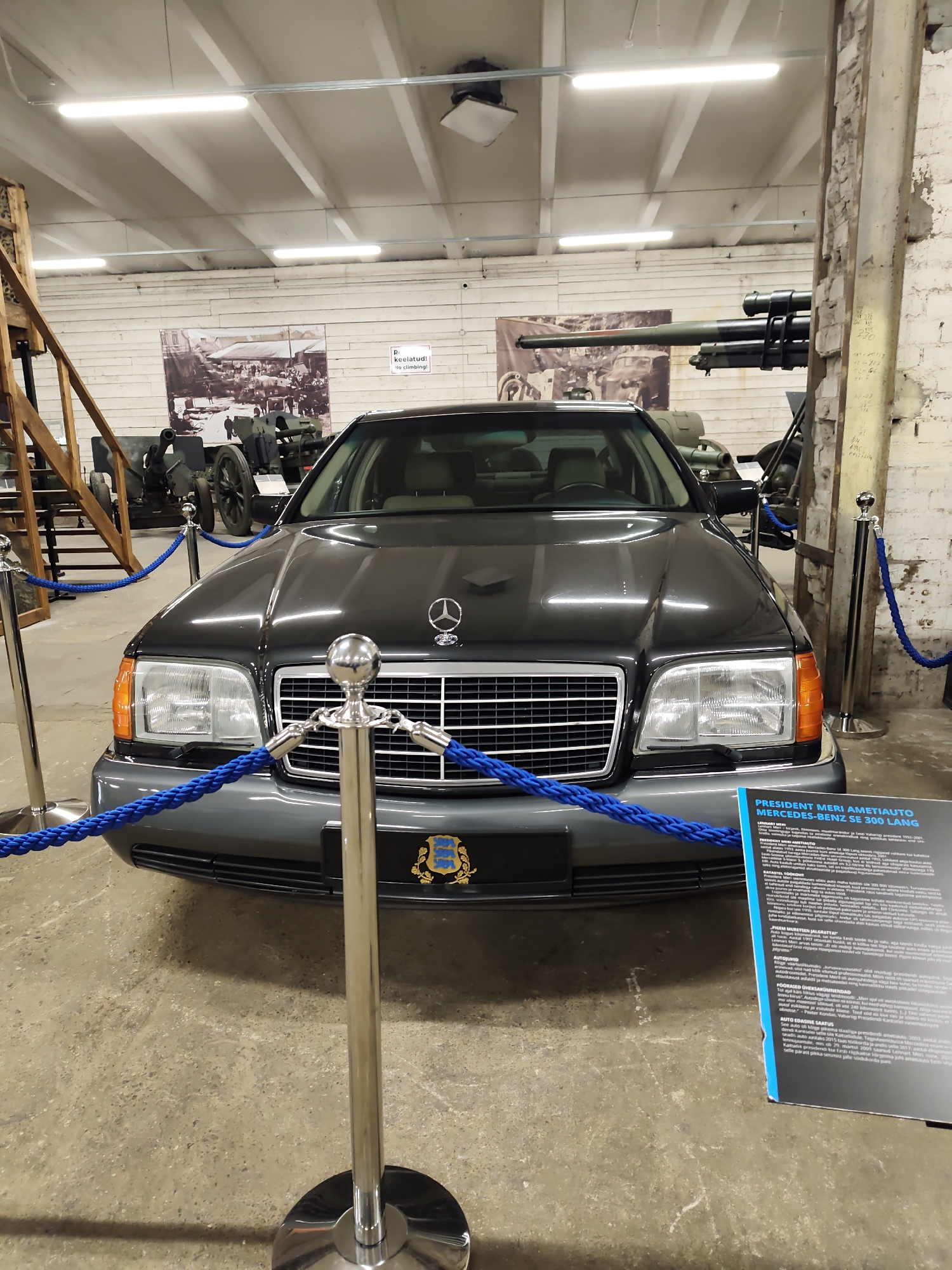 The image shows a dark-colored Mercedes-Benz sedan, identified by a plaque as "President Meri Ametiauto Mercedes-Benz SE 300 Lang," displayed in a museum or exhibition space. The car is positioned centrally in the frame, facing forward, with blue velvet ropes and stanchions cordoning it off from the public. The car has a chrome grille with a Mercedes-Benz logo and distinct headlights with orange turn signals. On the front of the car, below the grille, is a crest that appears to be the coat of arms of Estonia.
The background reveals an indoor environment with a concrete floor and a high ceiling with exposed beams and fluorescent lighting. To the left of the car, there is a wooden staircase and scaffolding, suggesting a historical or industrial setting. In the background, there are large black and white photographs or murals depicting historical scenes with people and military equipment. Further in the background, there are large artillery pieces, indicating a military museum. To the right of the car, a brick wall with some markings and what appears to be part of a gun barrel are visible. A blue information plaque with text in Estonian is placed in the lower right corner of the image. The overall impression is of a well-maintained vehicle presented as a historical artifact in a museum.