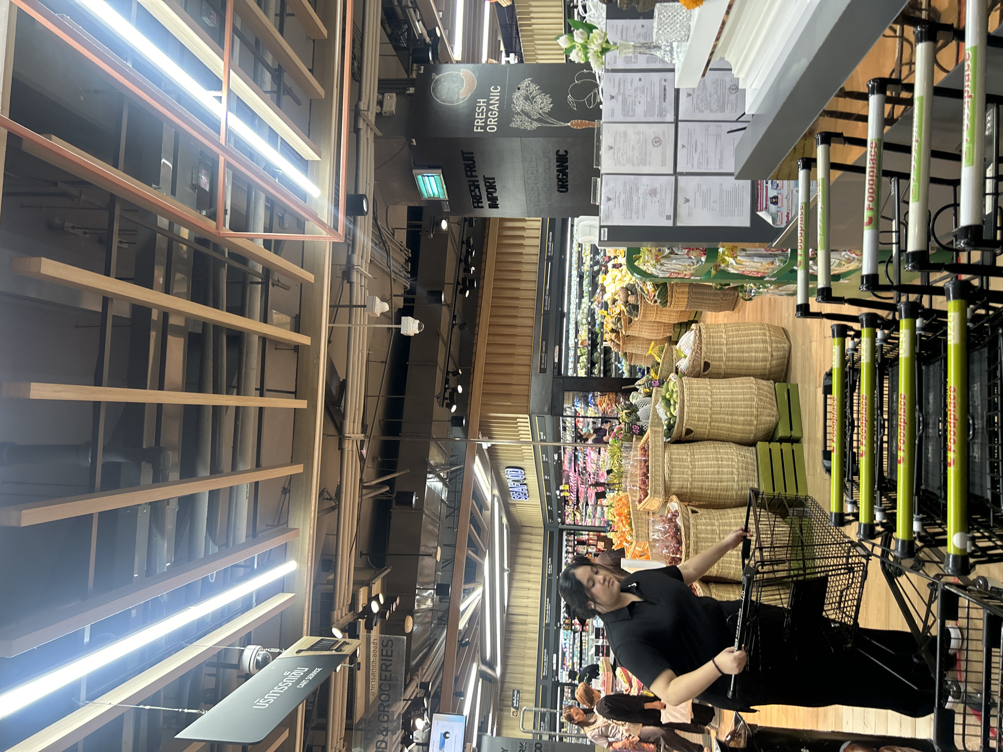 This is a medium-shot, eye-level interior photograph taken inside a supermarket in Bangkok, Thailand. The scene depicts a checkout area with multiple shopping carts lined up. A woman wearing a black polo shirt and black pants is pushing a shopping cart in the foreground. She is facing away from the camera and appears to be an employee of the supermarket. Behind her, there are rows of shopping carts with green handles, many of which have "Foodplace" branding on them.
In the background, a produce section is visible with various fruits and vegetables displayed in wicker baskets. Signs with Thai script and English text are present, including one that reads "FRESH FRUIT IMPORT" and another that says "FRESH ORGANIC" with an illustration of a carrot and vegetables. The ceiling has exposed wooden beams and fluorescent lighting. The overall environment suggests a well-lit, modern grocery store.