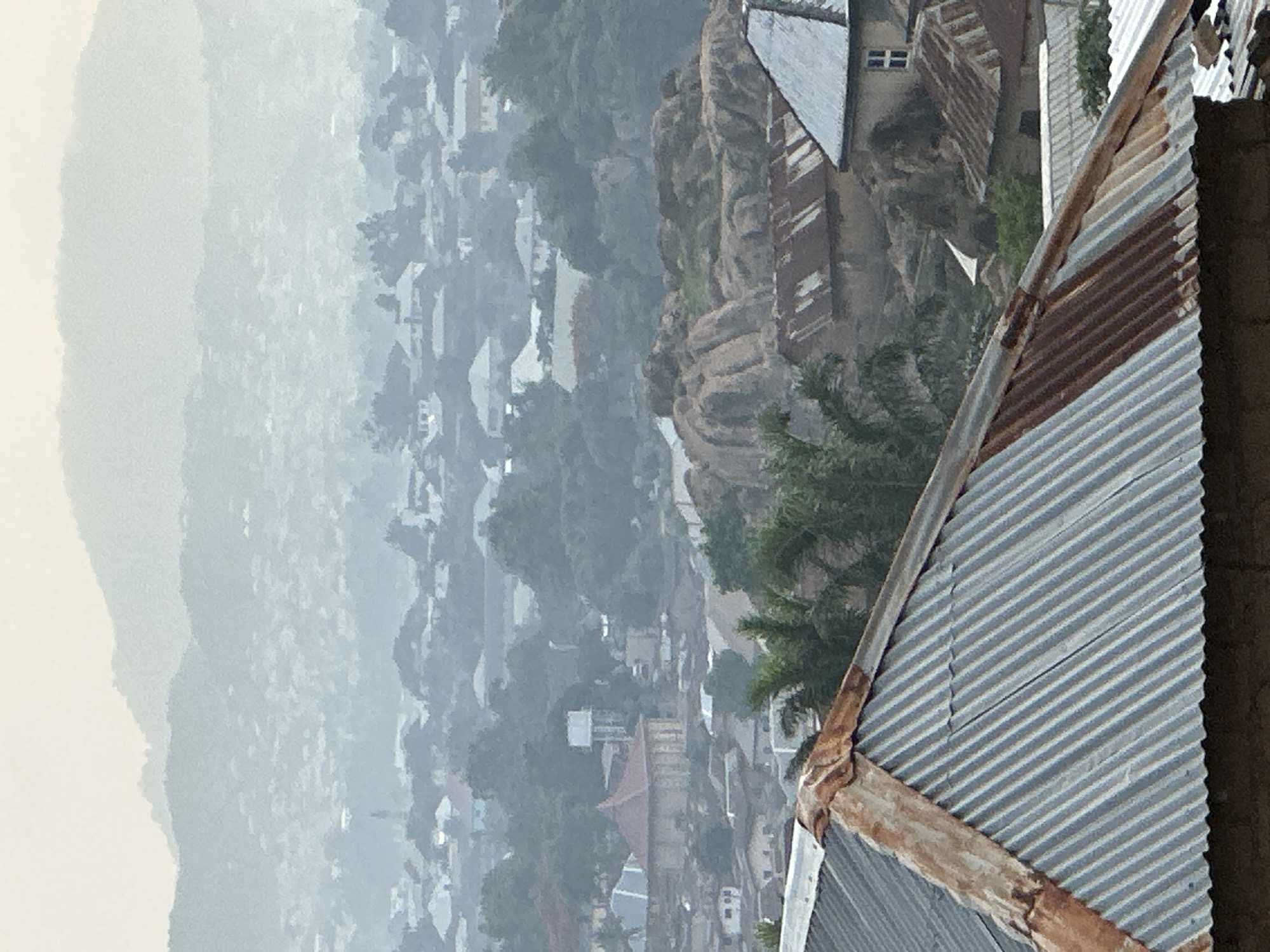 The image presents a panoramic view of a densely built-up area, likely a city or town in Jos, Nigeria, taken from a high vantage point looking down. In the immediate foreground, a weathered, corrugated metal roof is prominently featured, slanting diagonally across the right side of the frame. The roof shows signs of rust along its edges and seams, indicating age or exposure to elements.
Beyond the foreground roof, the landscape unfolds into a sprawling settlement characterized by numerous buildings, many of which also appear to have corrugated iron roofs. The urban area is interspersed with rocky outcrops and large boulders, suggesting a hilly or rugged terrain. Green foliage from trees is scattered throughout the settlement and on the rocky elevations, adding pockets of nature to the urban environment.
In the midground and background, the density of buildings continues, gradually becoming hazier and less distinct due to atmospheric conditions, possibly mist or dust. The distant horizon reveals layers of hazy mountains or hills, fading into the pale sky.
No people are visible in the image, and there is no specific activity taking place; it is a static landscape view. The lighting suggests it is daytime, with a soft, somewhat diffused light that could be characteristic of early morning, late afternoon, or a hazy day. No visible text is present in the media. The overall impression is that of an expansive urban setting nestled within a distinctive rocky landscape.