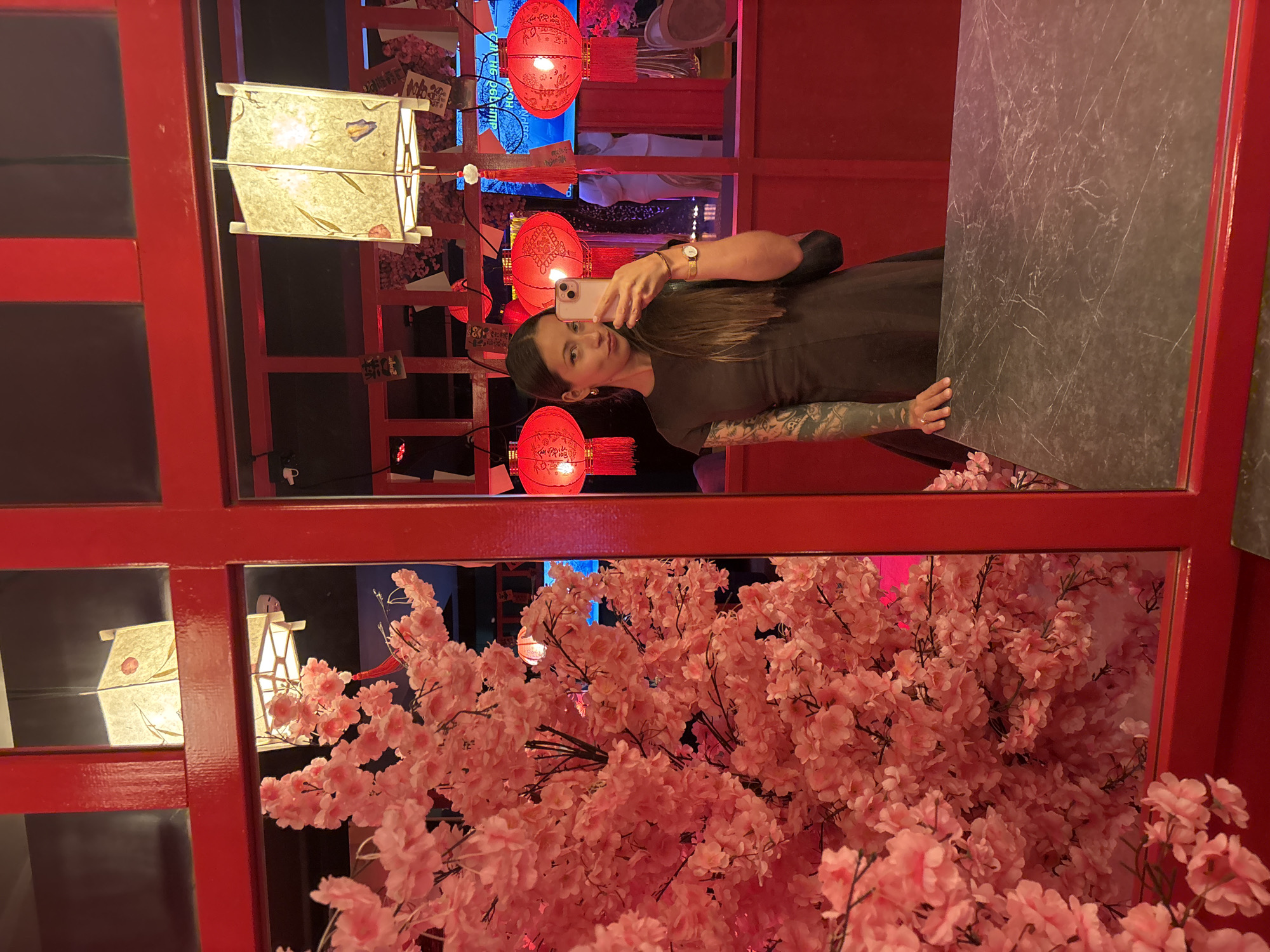 The image captures the interior of Red. Wok. Cafe & Bar, located in Saint Petersburg, Russia. A woman is positioned to take a mirror selfie, holding a light-colored smartphone, likely an iPhone, in her left hand to capture her reflection. She has long, dark, straight hair, wears a dark short-sleeved dress, and her left arm is adorned with extensive tattoos. A gold-toned watch is visible on her right wrist, which rests on a dark grey table with white marble-like veining in the lower right foreground.
The environment is characterized by a vibrant red and pink color scheme. Bright red architectural framing, possibly window or room dividers, dissects the scene. The left side of the image is filled with dense artificial pink cherry blossoms, arranged as a significant floral display. Overhead, square paper lanterns with delicate botanical designs, including leaves, flowers, and butterflies, provide ambient lighting. In the reflection behind the woman, multiple large, round red paper lanterns with visible Chinese characters and red tassels hang in the background. Small white paper tags are suspended from a red grid structure visible between the lanterns. A blurry screen displaying text and the faint reflection of another individual in light attire can be discerned in the deep background of the mirror.
